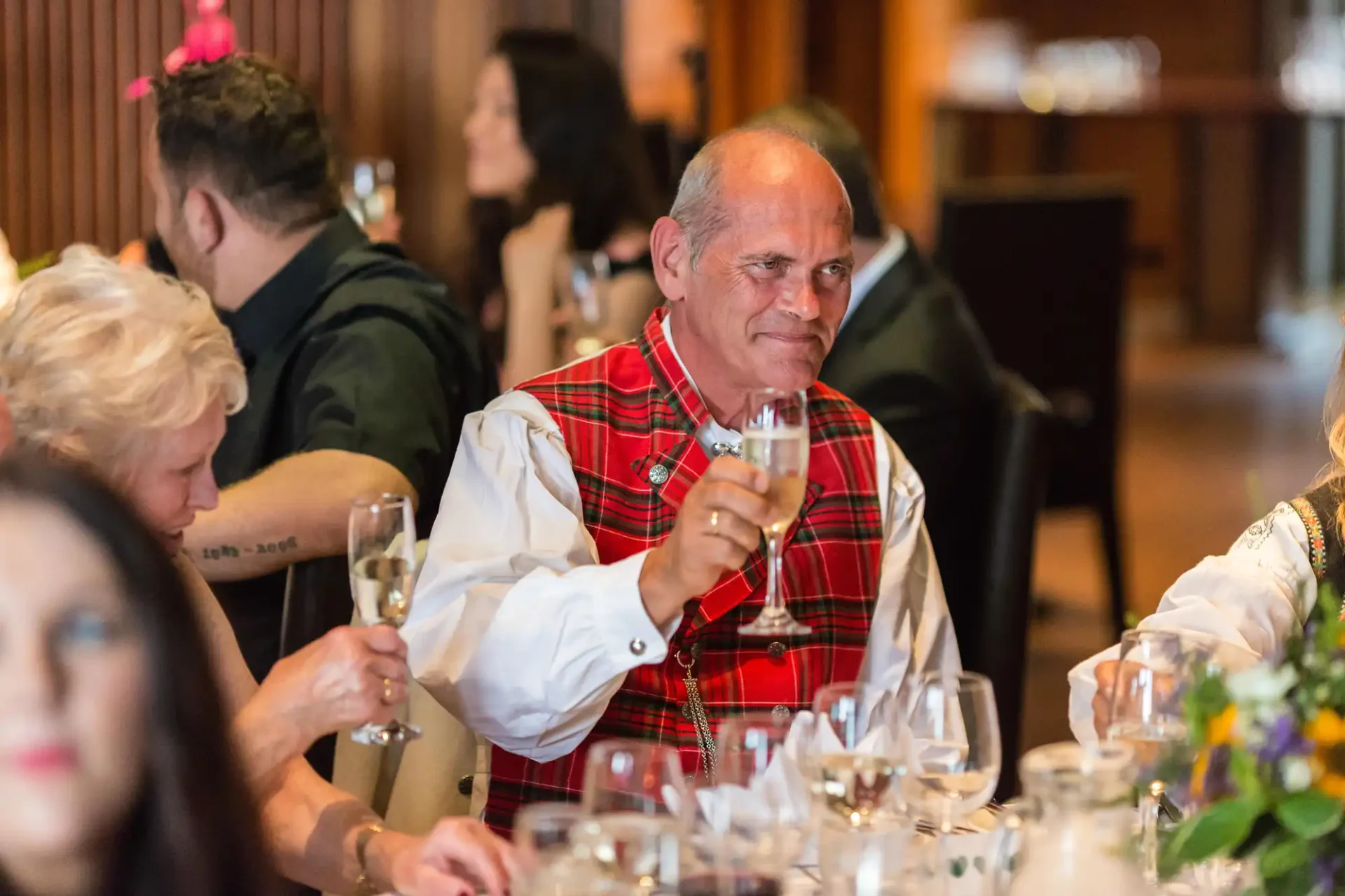A man in a tartan plaid shirt raising a glass at a social event with guests sitting at tables in the background.