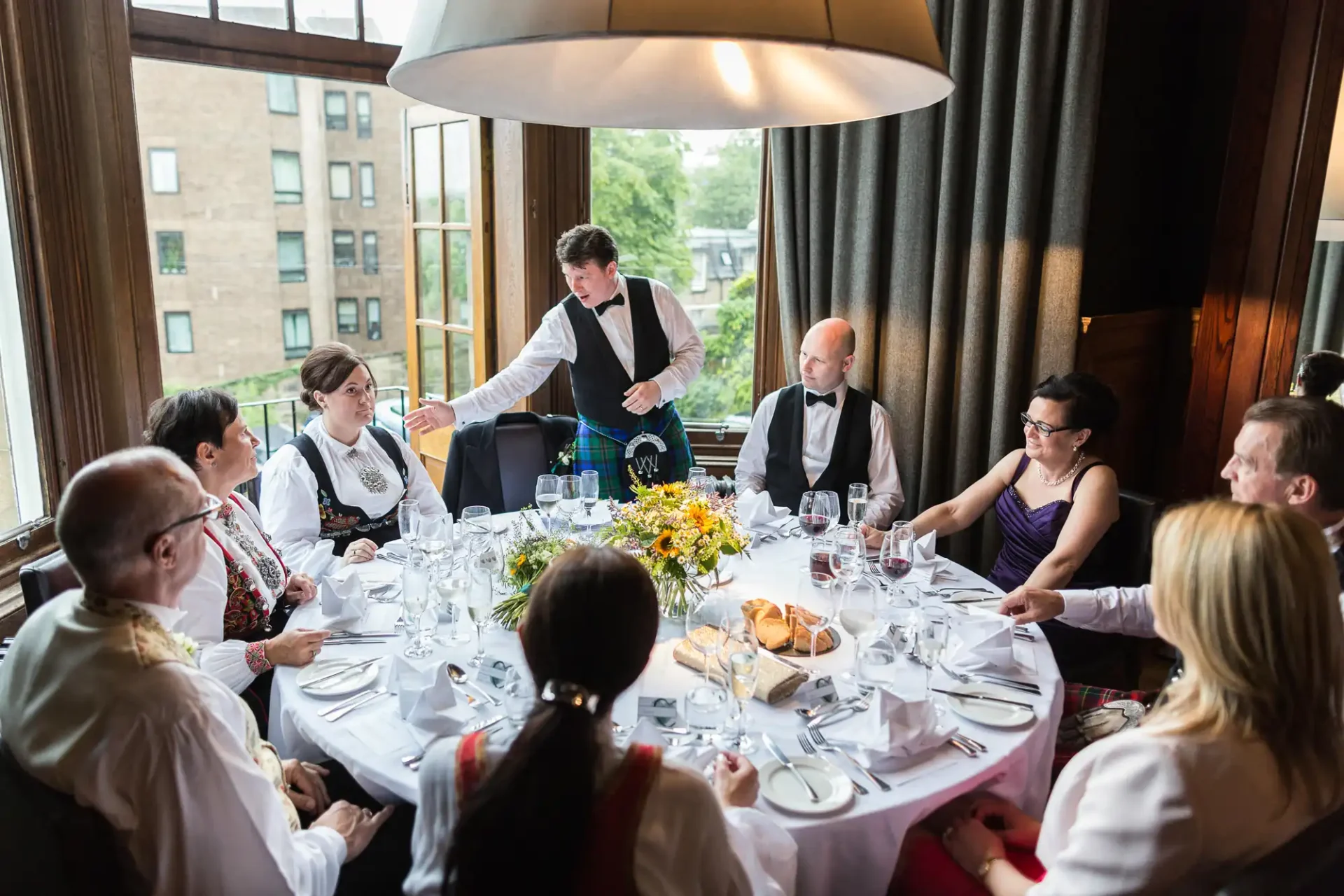 A waiter pours wine for a guest at a formal dining event surrounded by elegantly dressed people seated around a table adorned with flowers.
