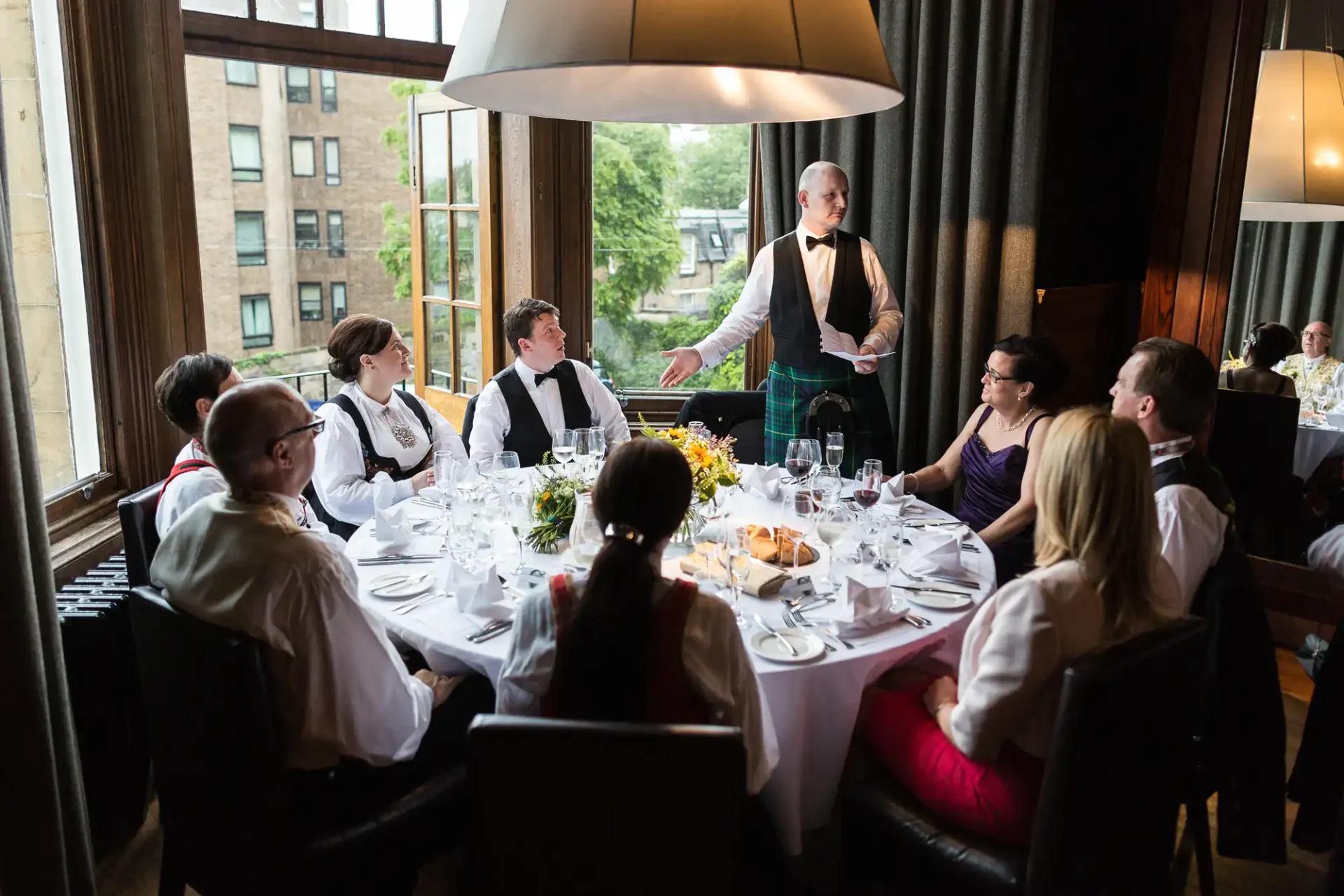 A waiter in a formal outfit addresses a group of guests seated around a dining table in an upscale restaurant.