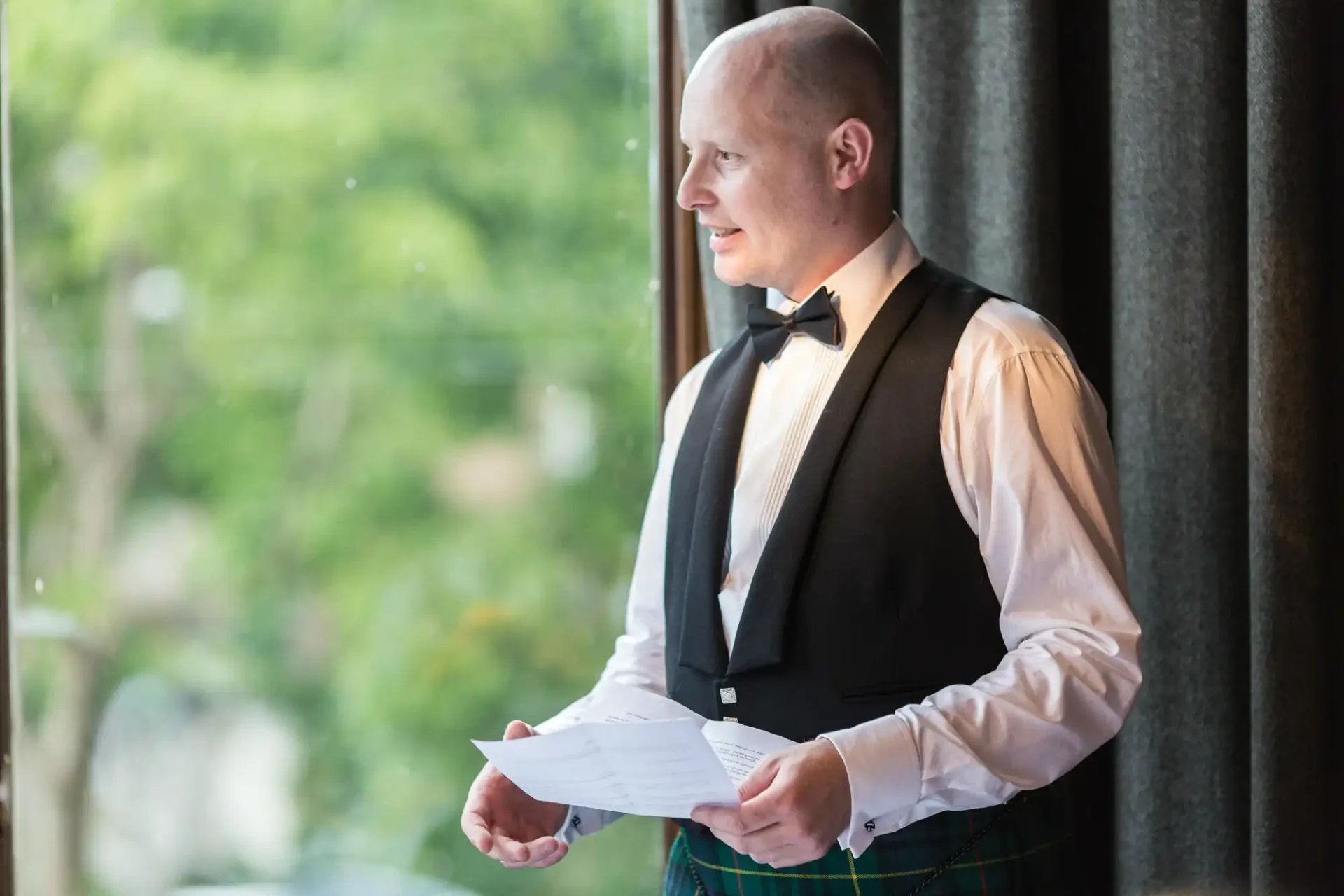 A bald man in a kilt and formal vest, holding papers and looking out a window.
