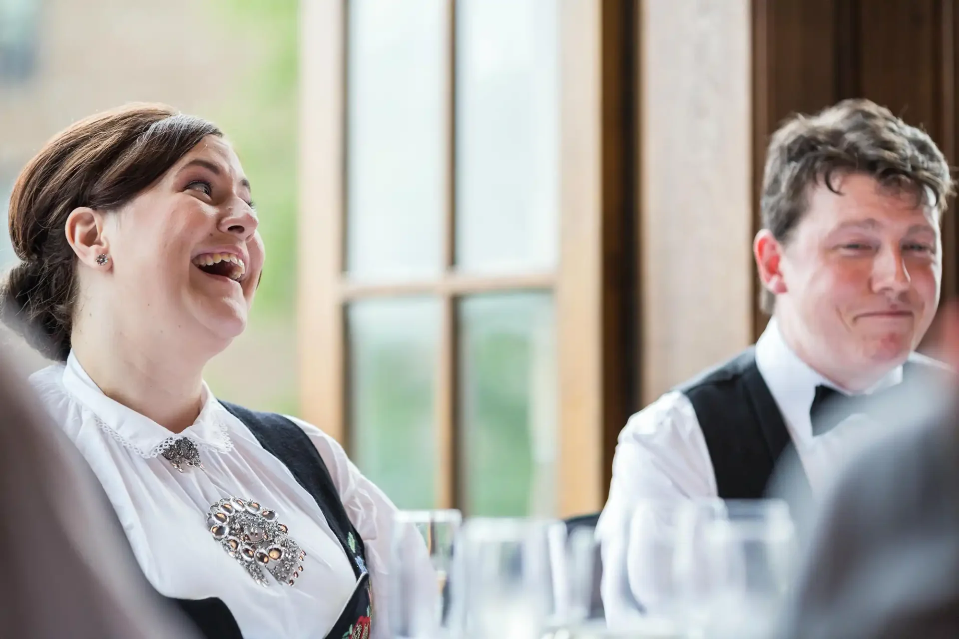 Woman in embroidered blouse laughing joyfully at a dining table beside a man in a tuxedo who is smiling.