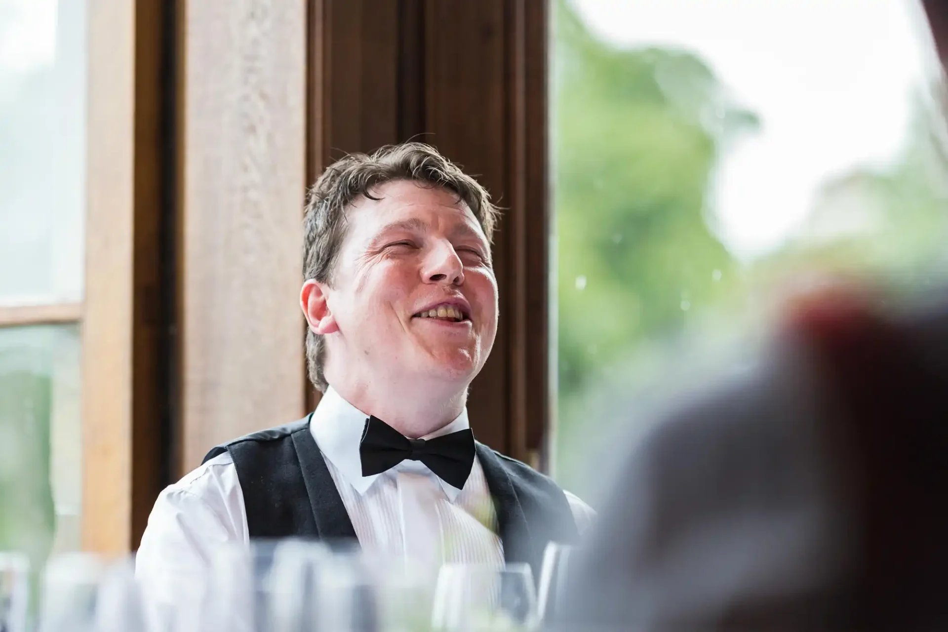 Man in a tuxedo with a bow tie laughing joyfully at a social event, seated indoors with large windows in the background.