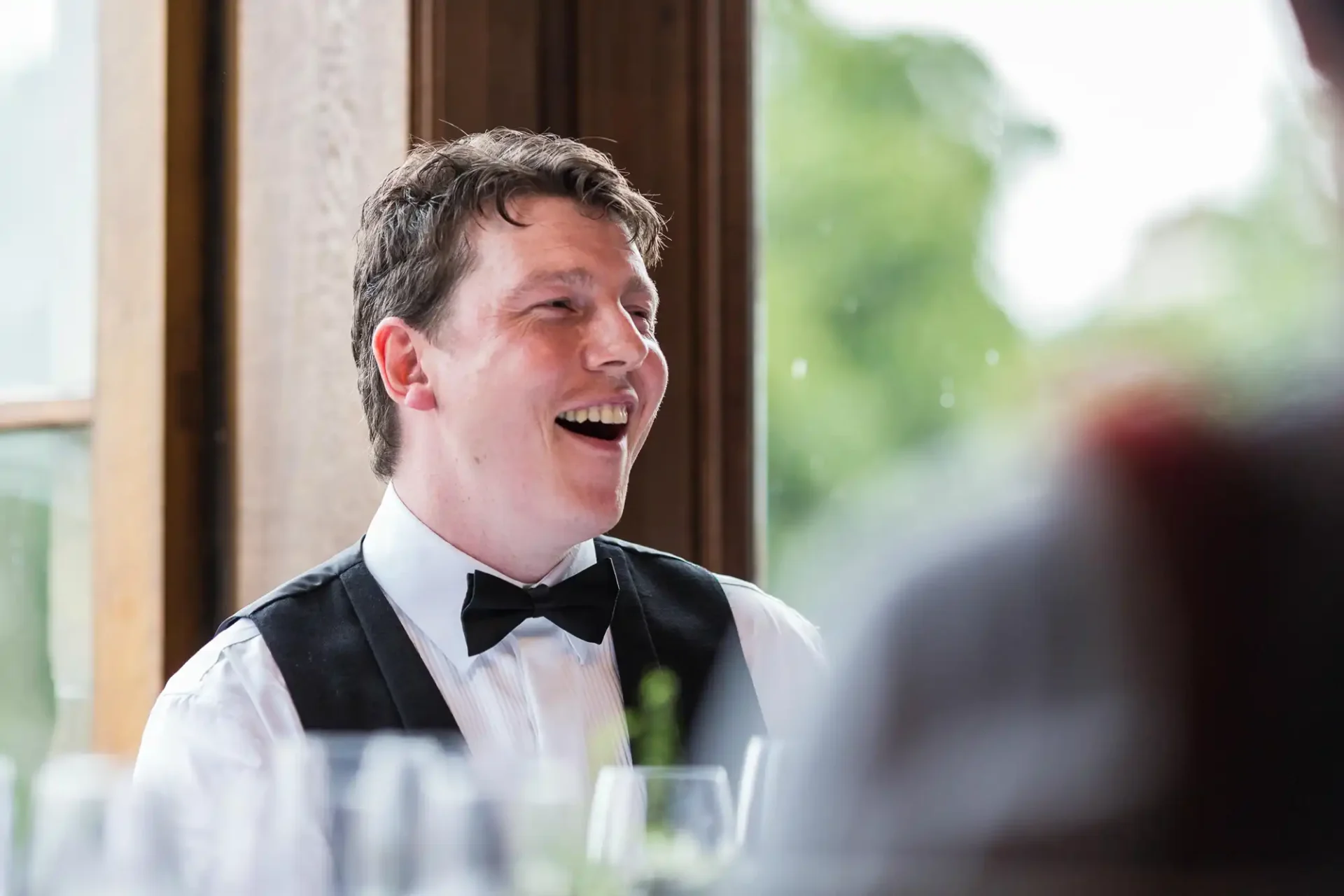 Man in a tuxedo smiling and laughing during a conversation at a formal event, seated indoors near a window.