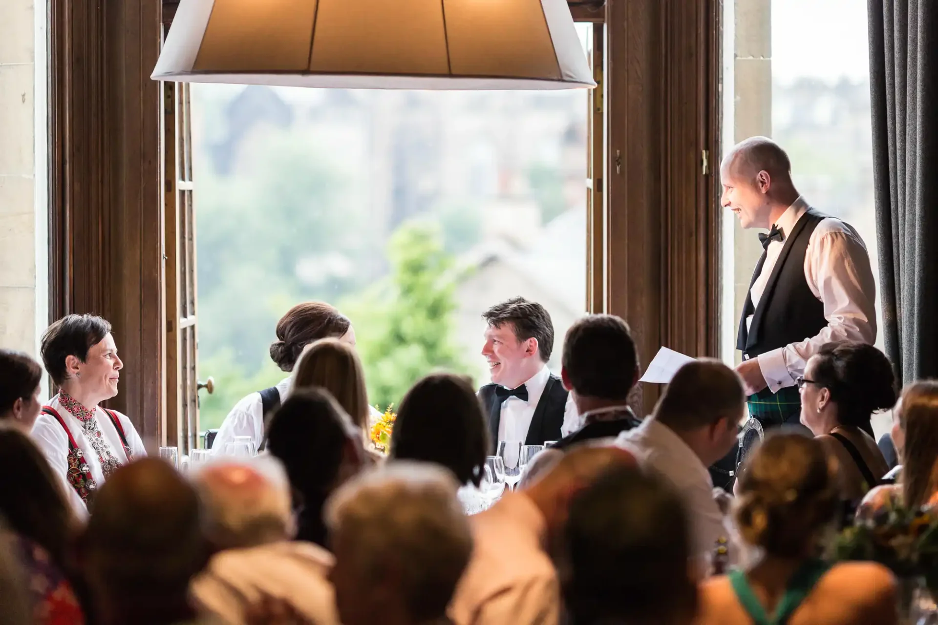 A man stands speaking to guests at a wedding reception inside a room with large windows overlooking a scenic view.