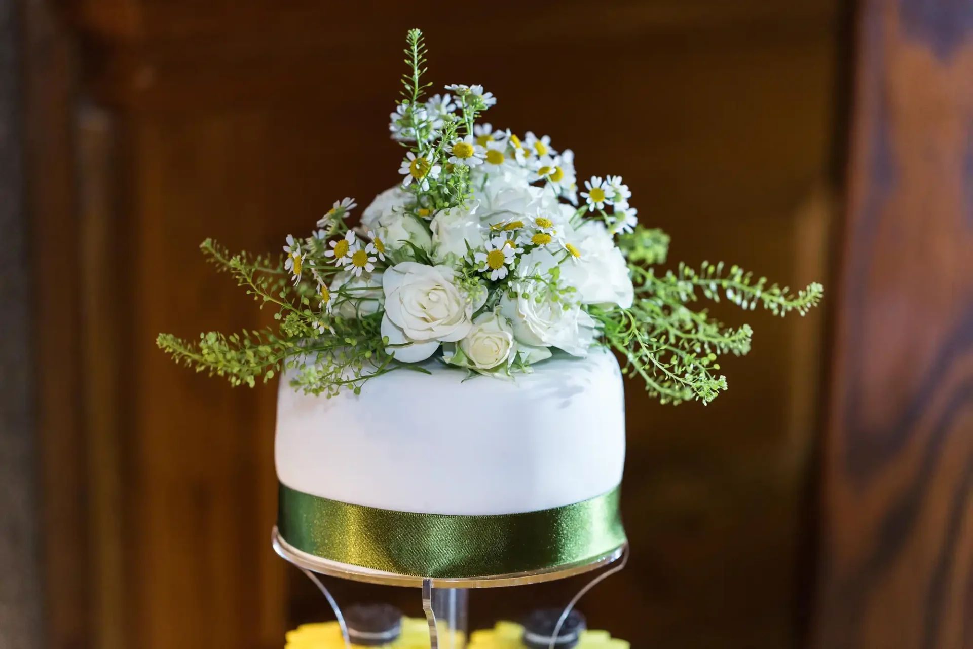 A white wedding cake decorated with fresh white roses and wildflowers, topped with greenery on a cake stand.