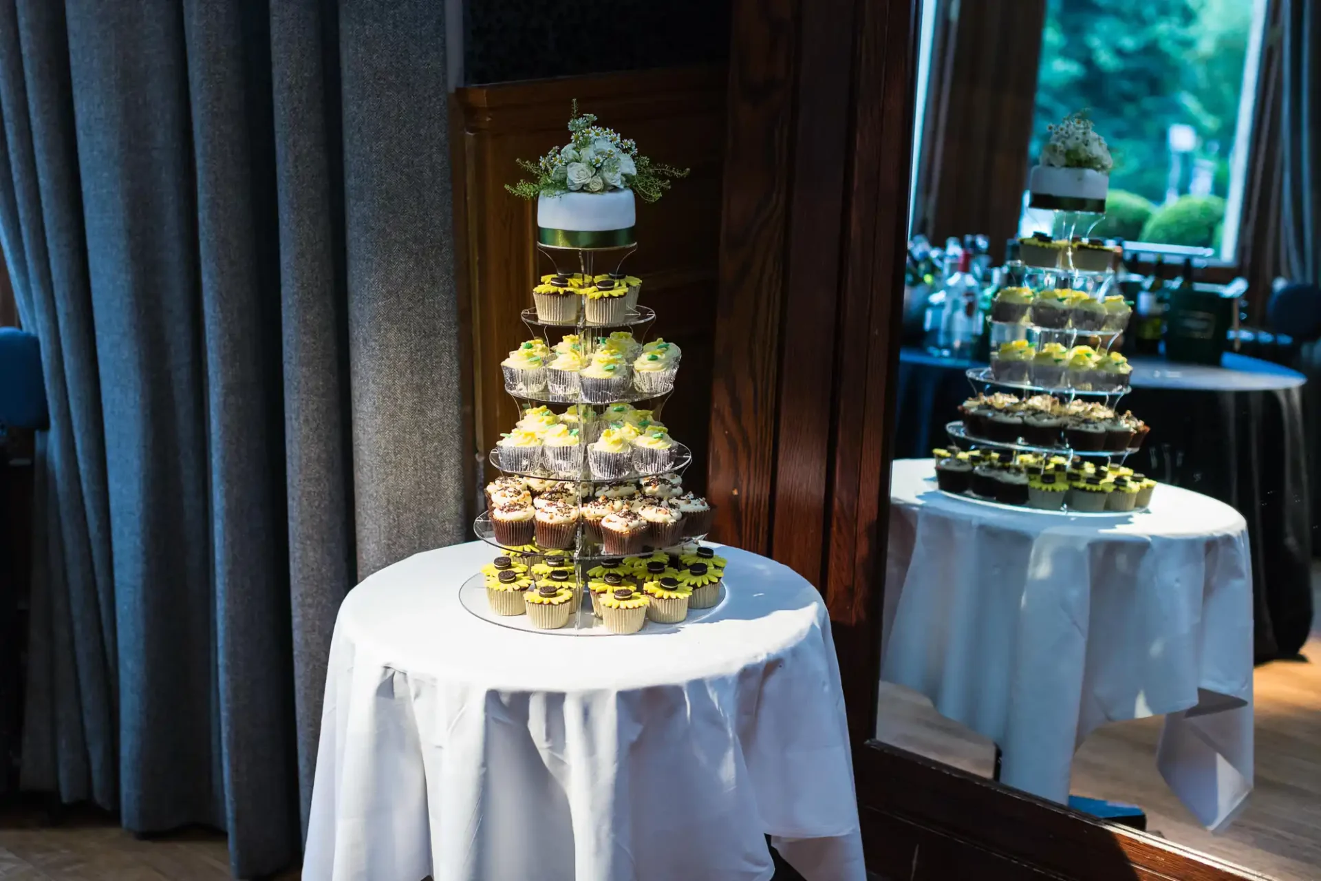 Tiers of assorted cupcakes on white cloth-covered tables, each topped with a floral arrangement, reflected in a large mirror in a dimly lit room.
