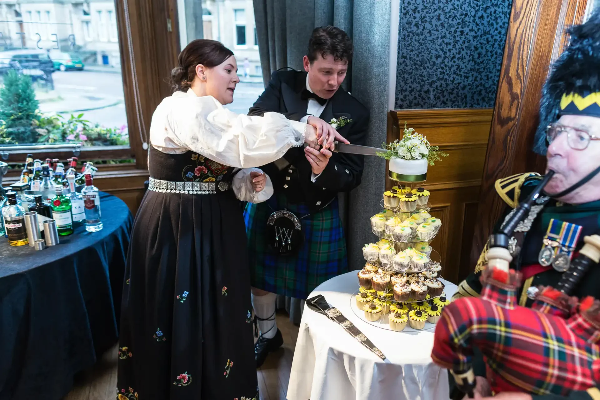 A couple in traditional scottish attire cuts a wedding cake while a bagpiper in a tartan kilt watches at an indoor reception.