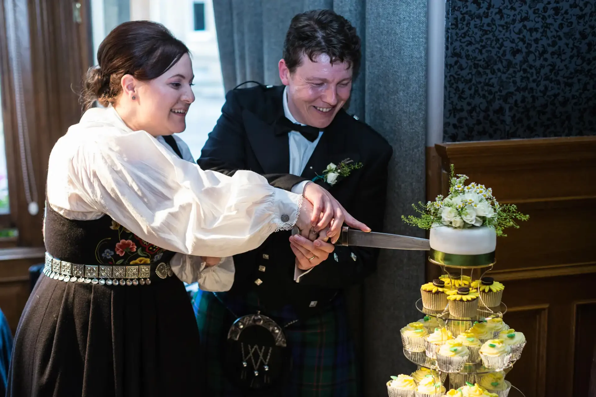 A bride and groom in traditional scottish attire smiling as they jointly cut a wedding cake with a sword.