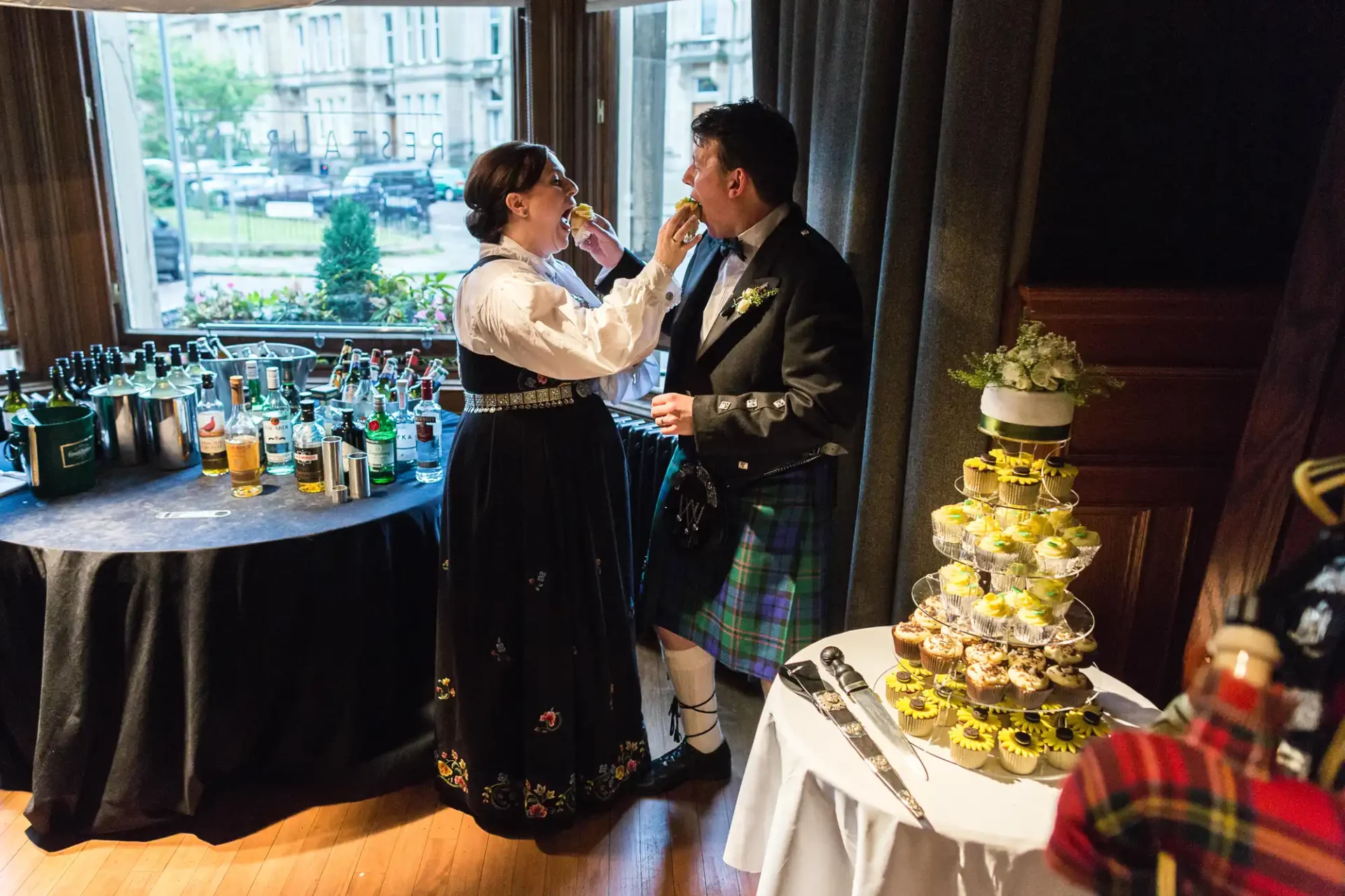 A couple in traditional scottish attire share an intimate moment at an indoor event, surrounded by a buffet and drinks table, with a tiered cupcake stand in the foreground.