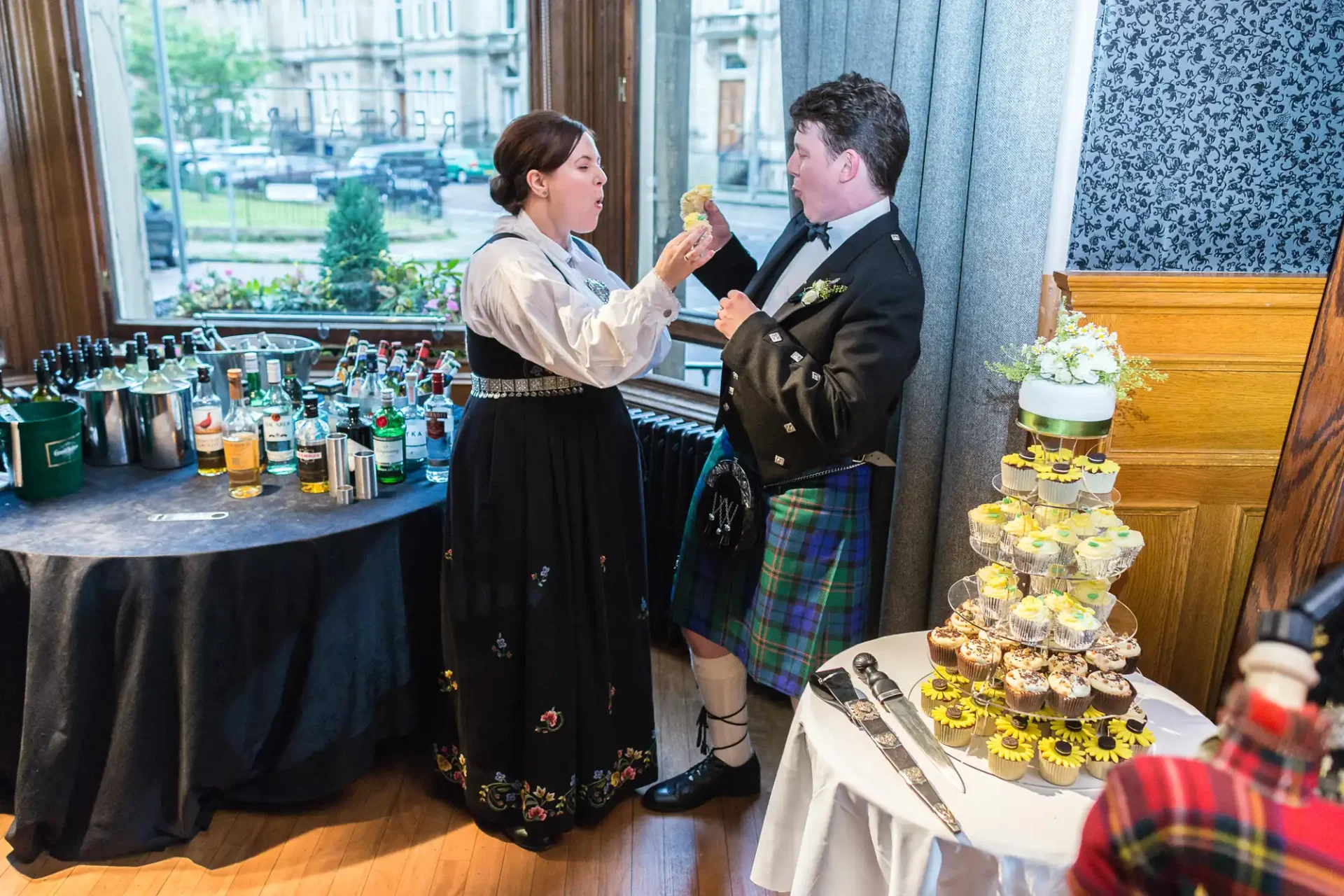 Two people in traditional scottish attire conversing beside a drinks table and cupcake stand at an indoor event.