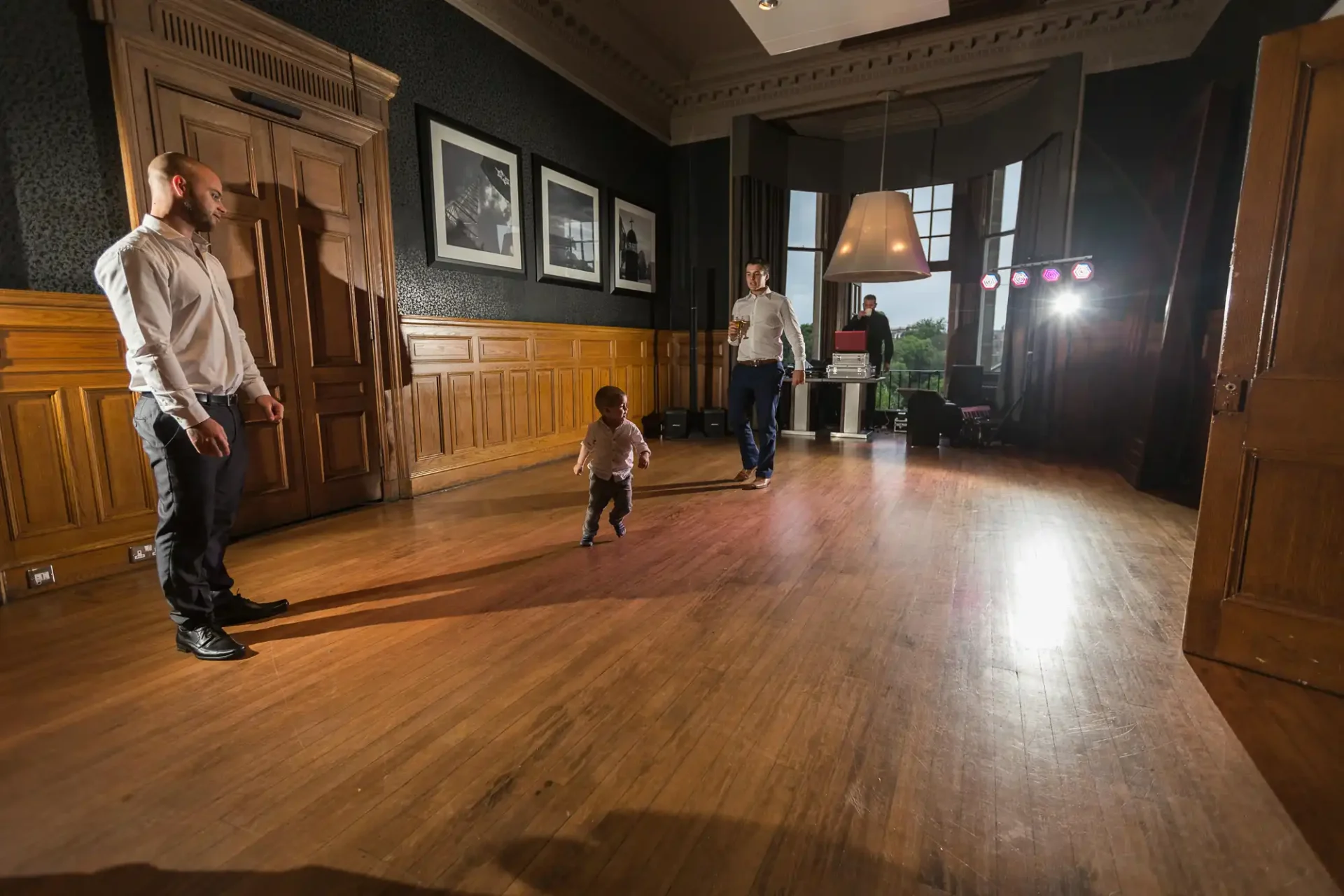 A toddler walks across a wooden-floored room towards two men standing near the entrance, with framed photographs on the walls.