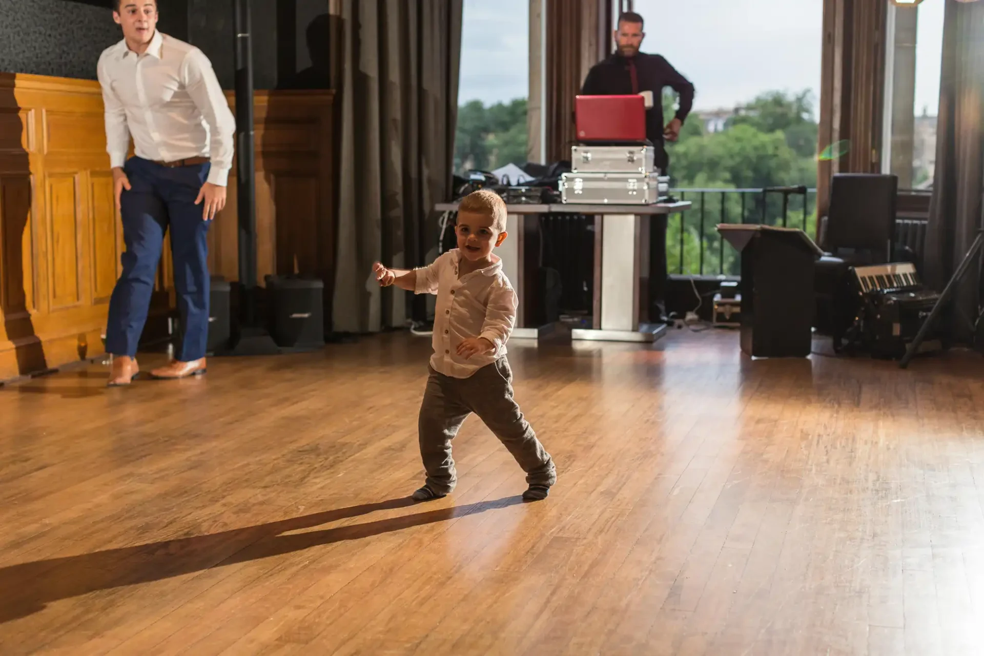 A young boy dances enthusiastically on a wooden floor at a social event, with a dj and another guest visible in the background.