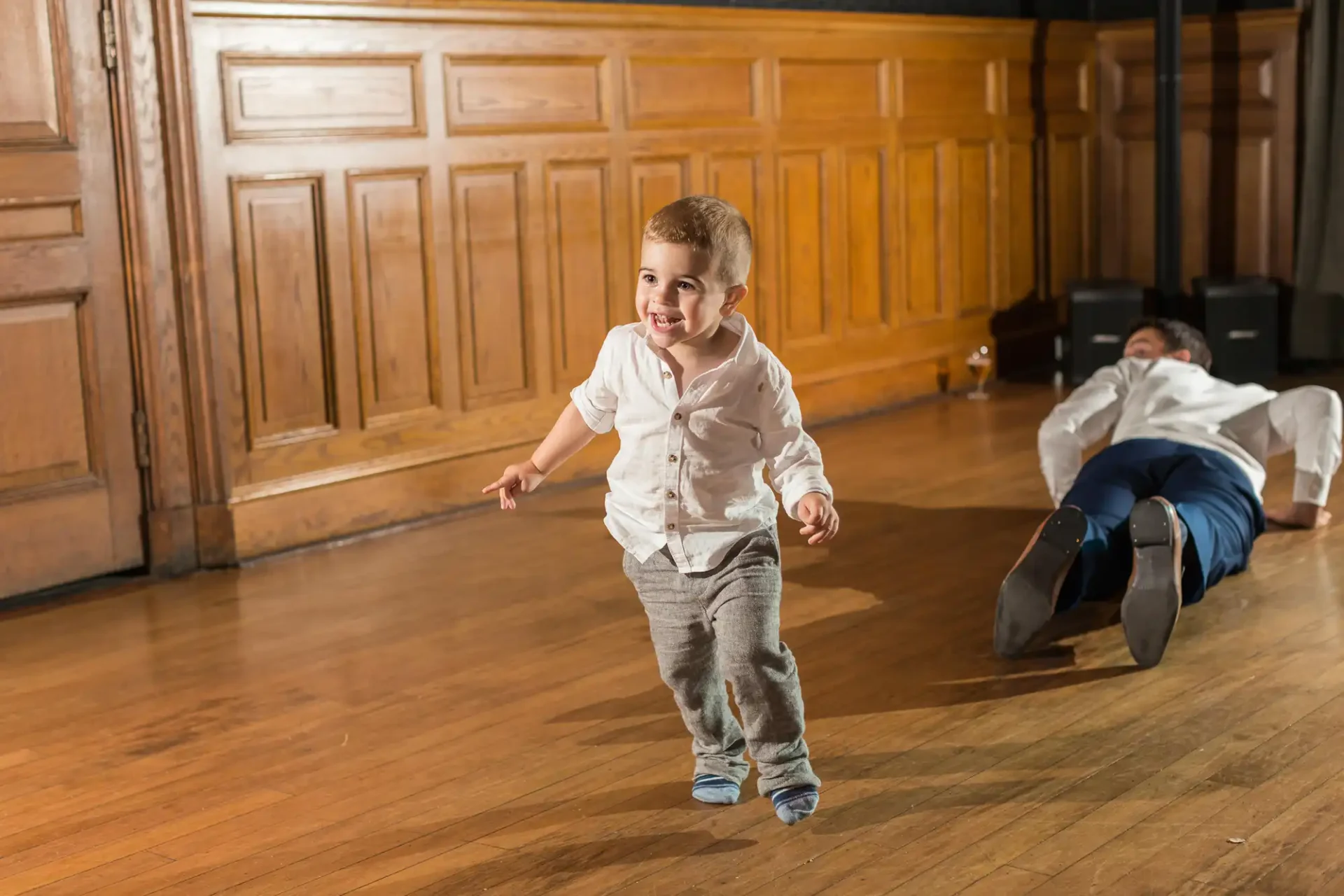 A young boy in a white shirt and jeans running joyfully in a wooden-paneled hall, with another child lying on the floor in the background.