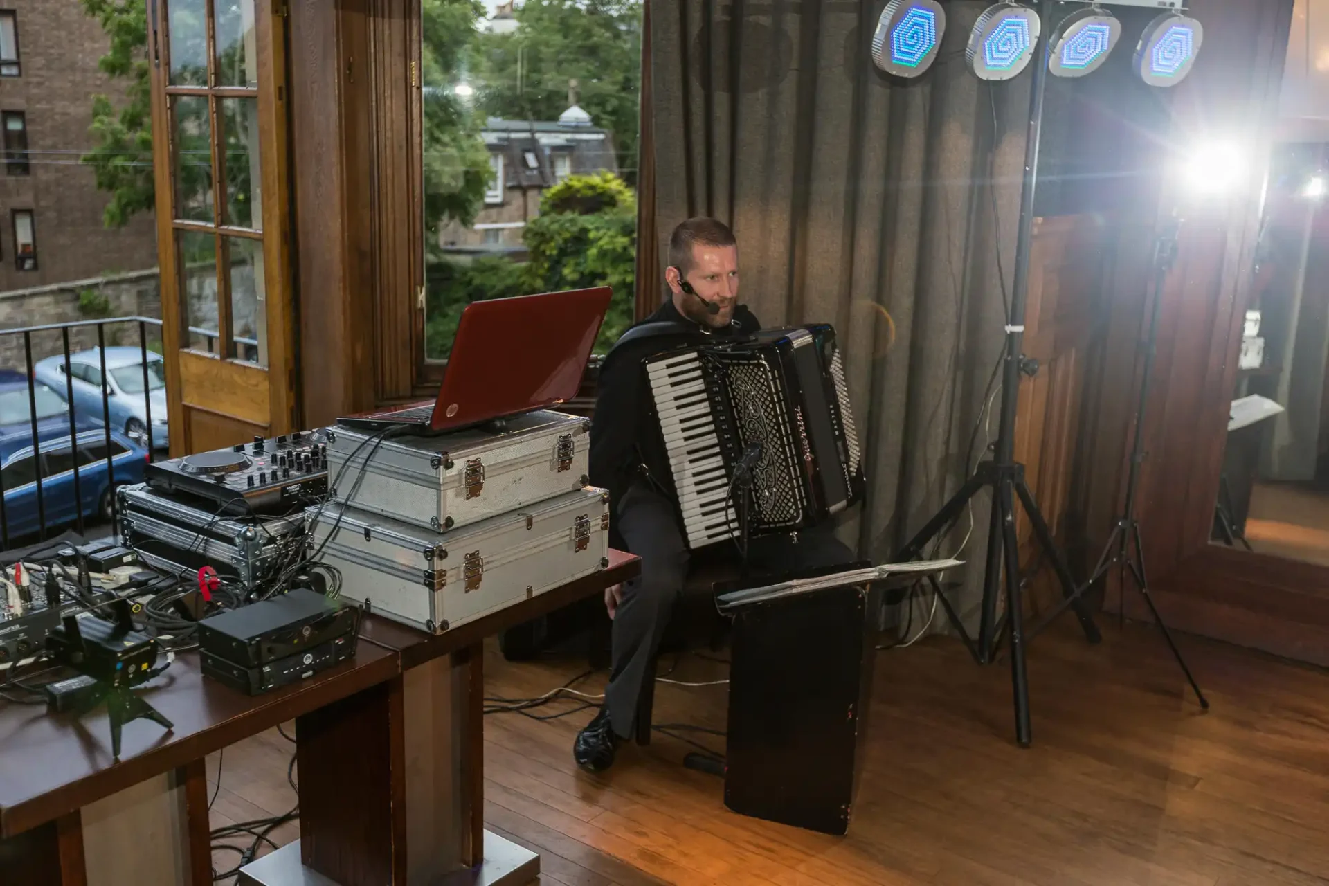 A man playing an accordion next to a table with sound equipment in a room with large windows overlooking trees.