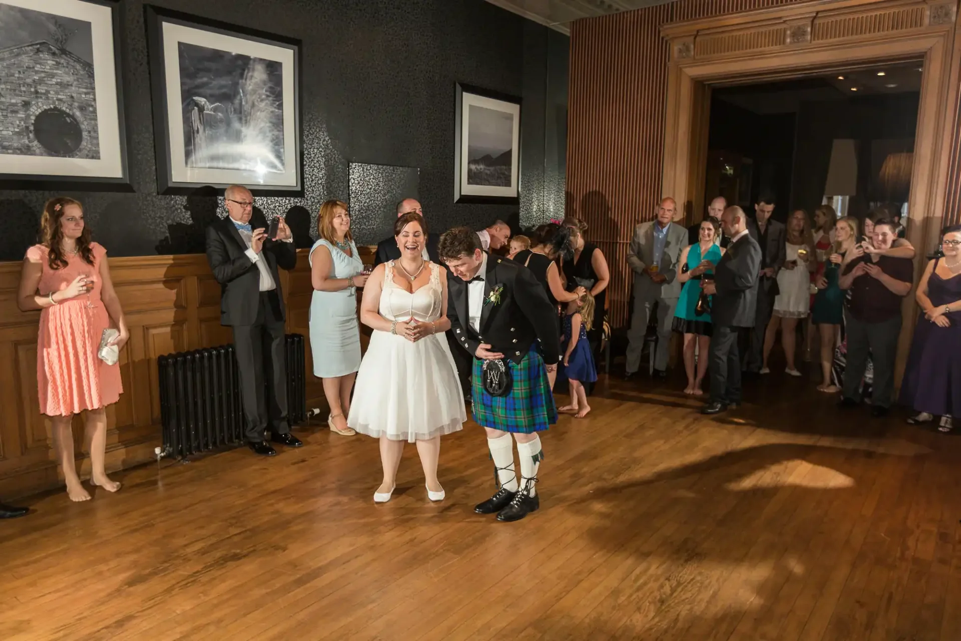 A bride and groom laugh together as they walk down the aisle, surrounded by guests in a warmly lit room with elegant decor. the groom wears a traditional kilt.