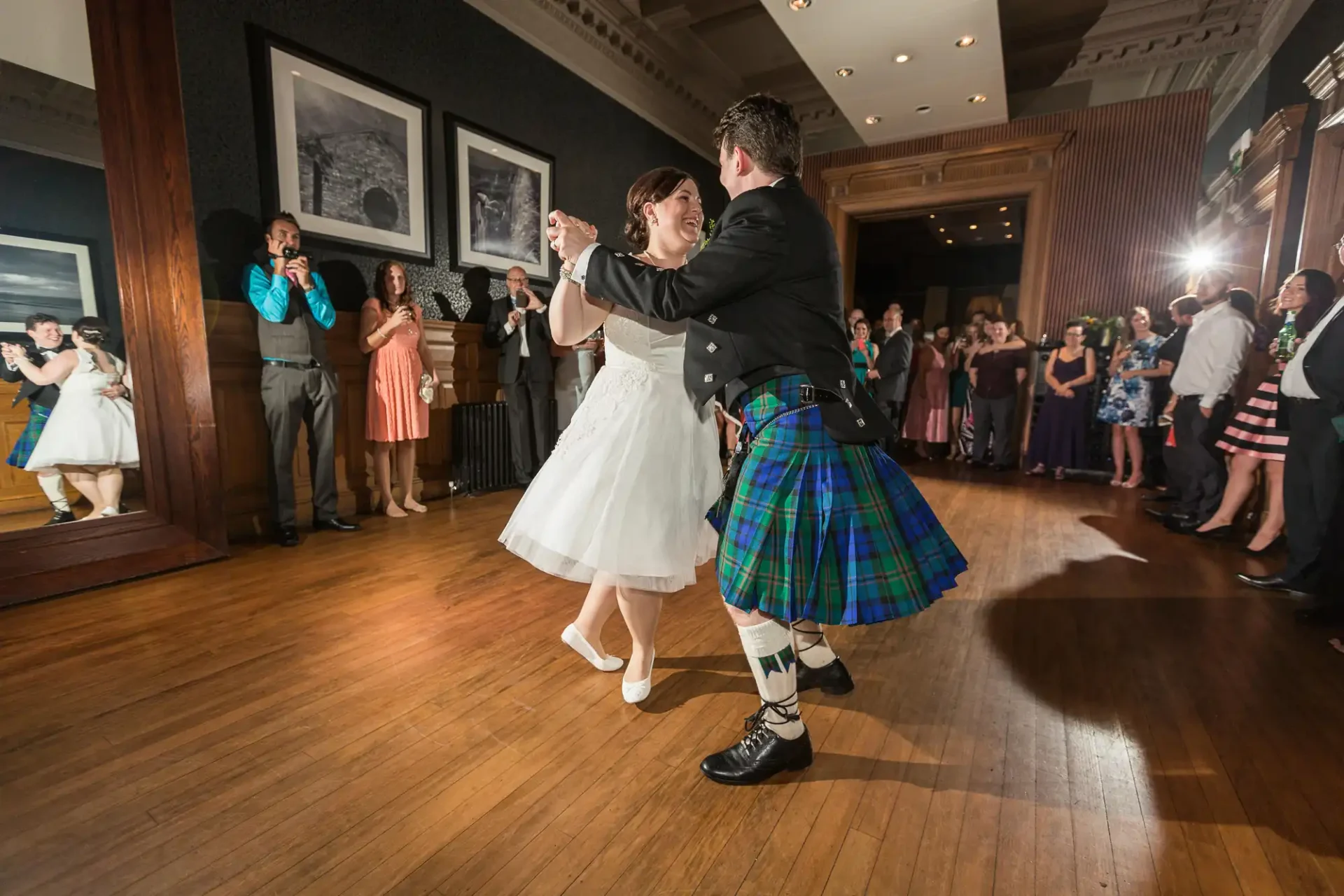 A joyful bride and groom dancing in a ballroom, surrounded by guests. the groom wears a kilt and the bride a short wedding dress.