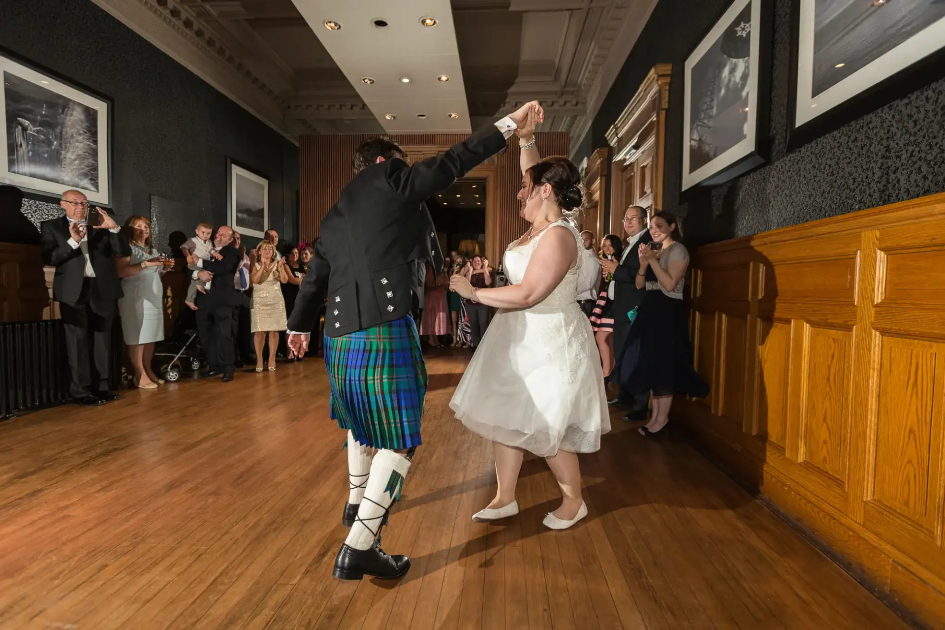 A bride and groom dancing joyously, the groom in a kilt, surrounded by applauding guests in an elegant hall.