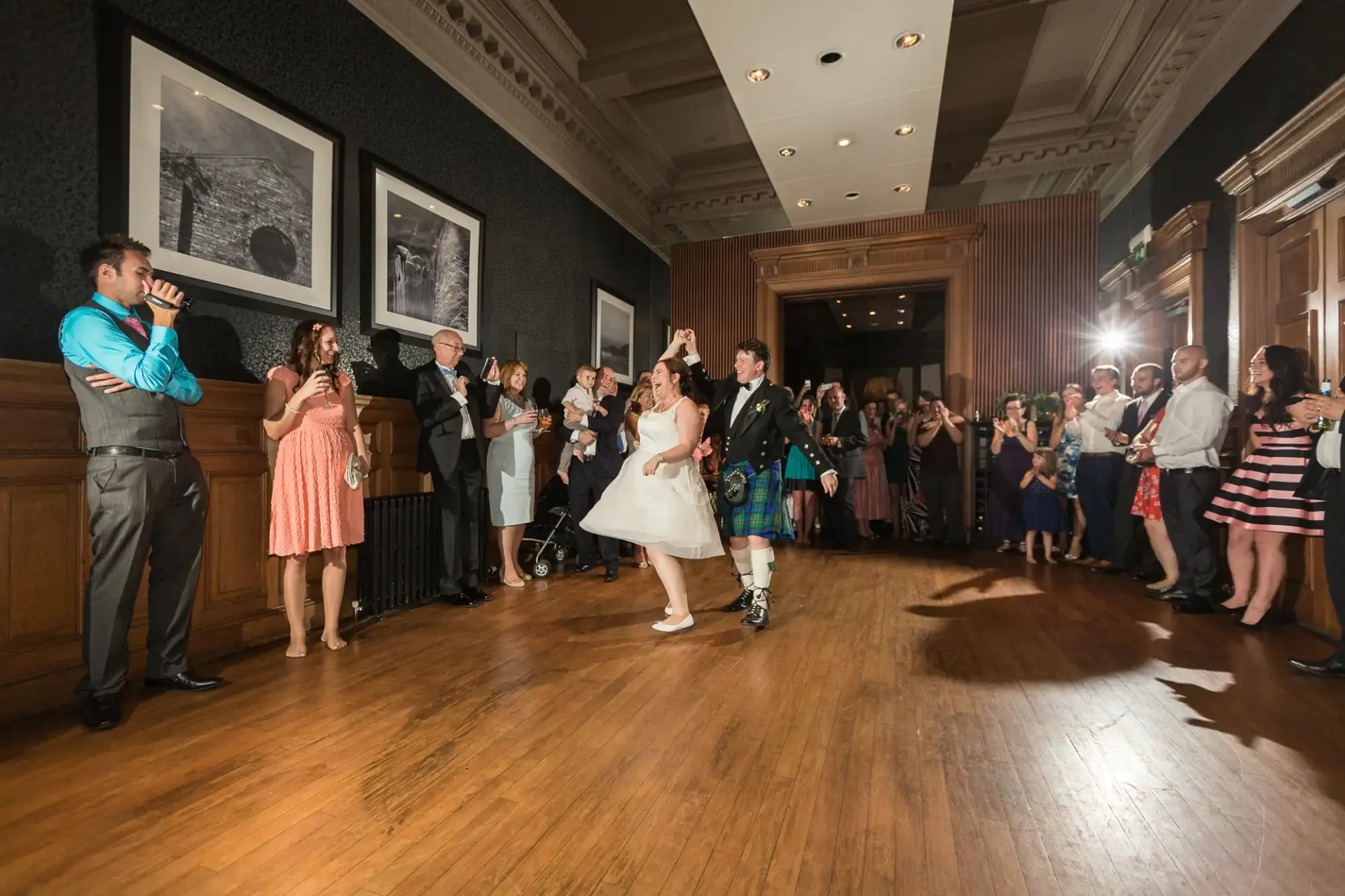 A bride and groom dancing joyfully in a ballroom surrounded by cheering guests and a photographer capturing the moment.