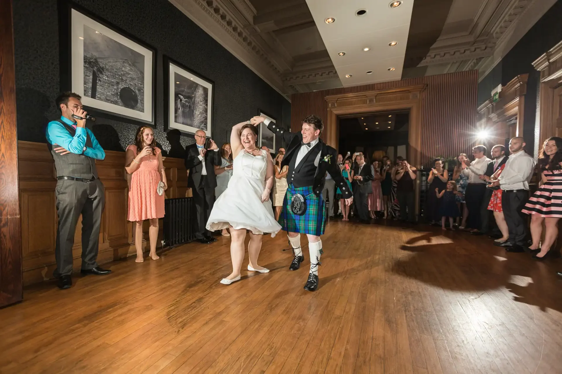 Bride and groom joyfully dancing in a hall with onlookers, the groom wearing a kilt and the bride in a short wedding dress.