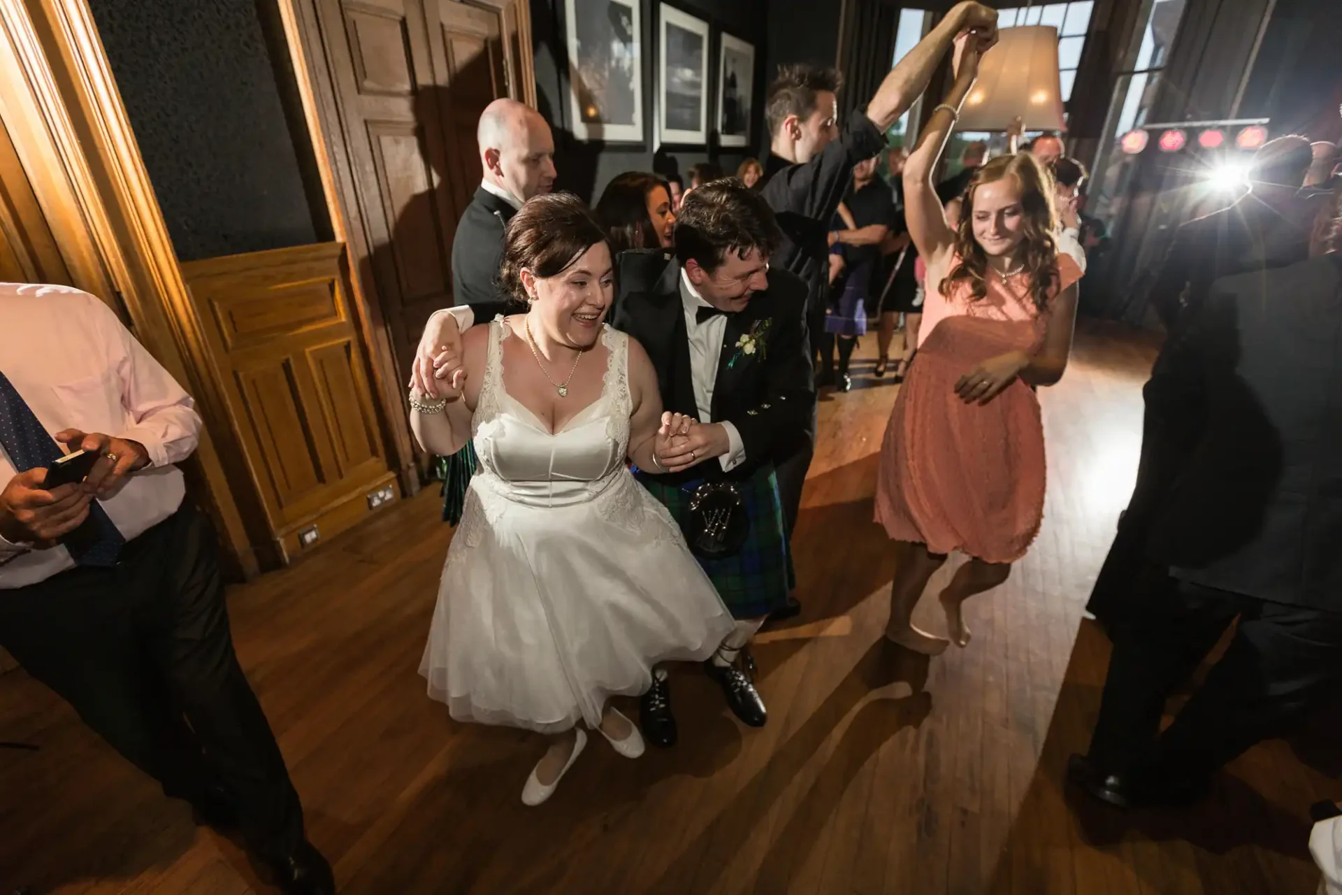 A bride in a white dress and a groom in a kilt dancing joyously among guests in a warmly lit room at a wedding reception.