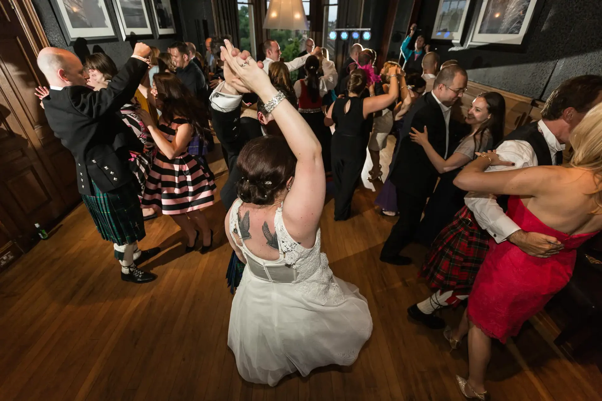 A bride in a white dress dancing with guests in a lively ballroom, some wearing kilts, under warm lighting.