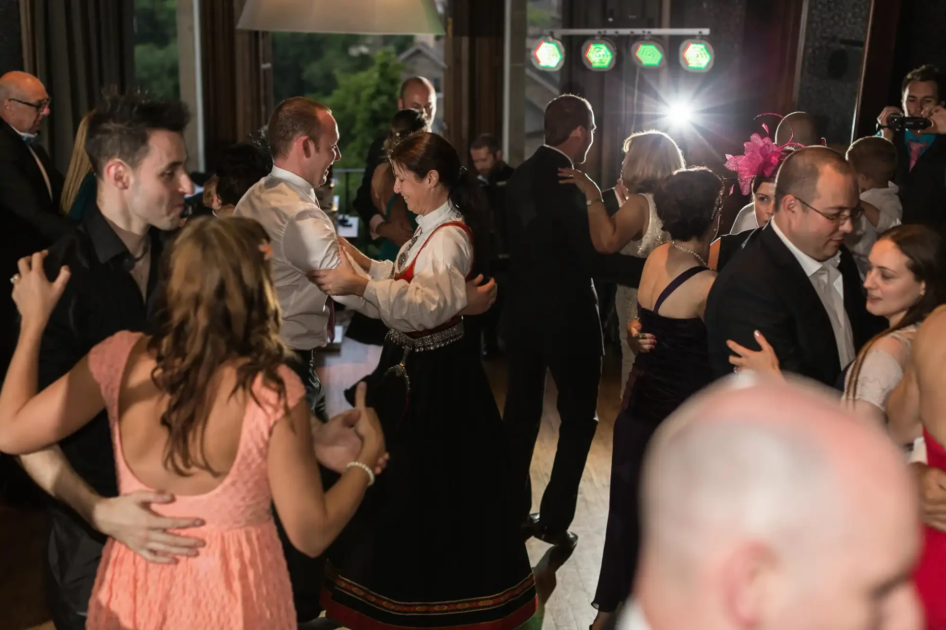 Guests dancing at a wedding reception, some in traditional attire, with colorful lights in the background.