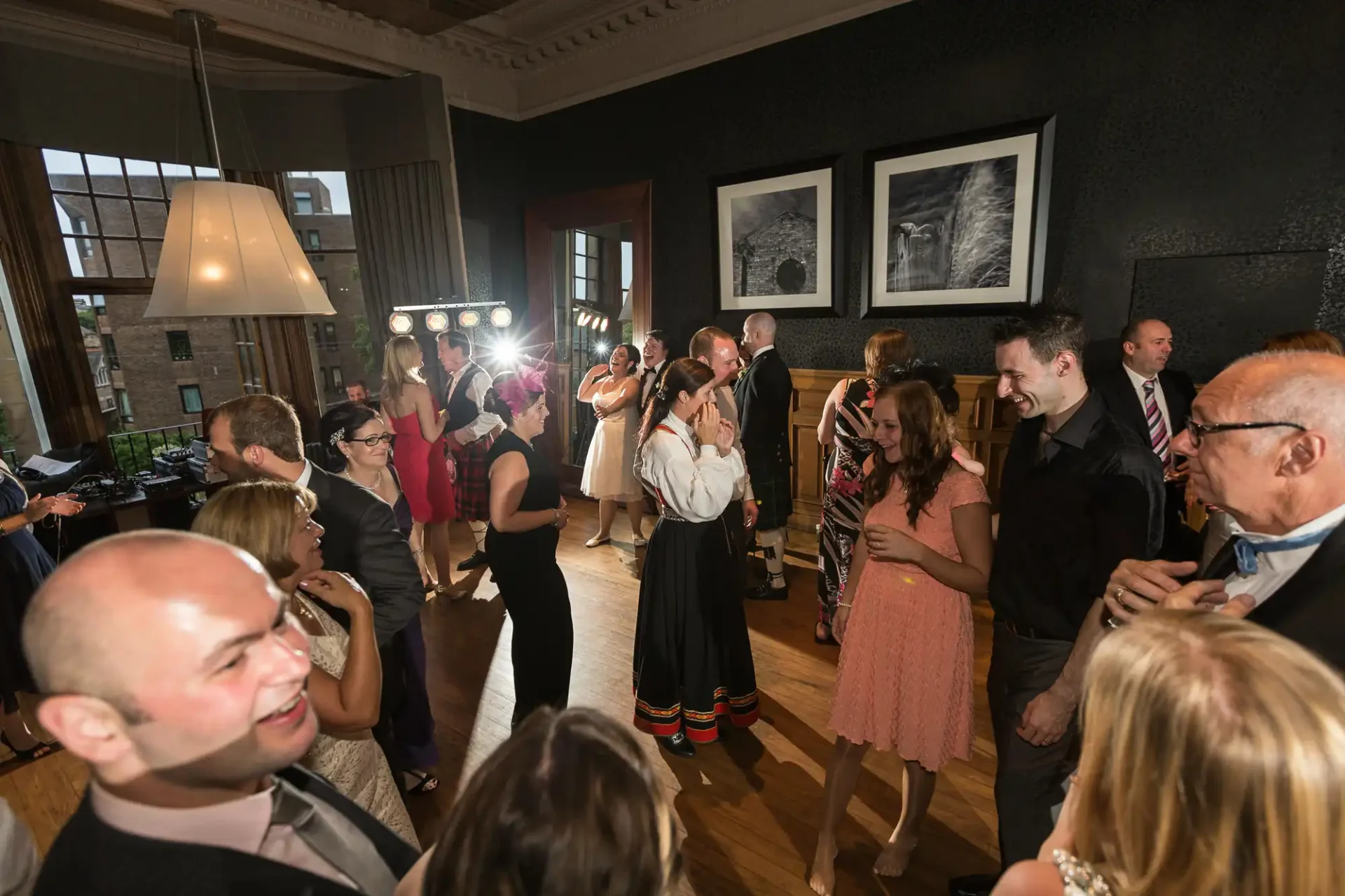 Guests in formal attire enjoy a lively indoor party with dancing and socializing in a room decorated with framed photographs.