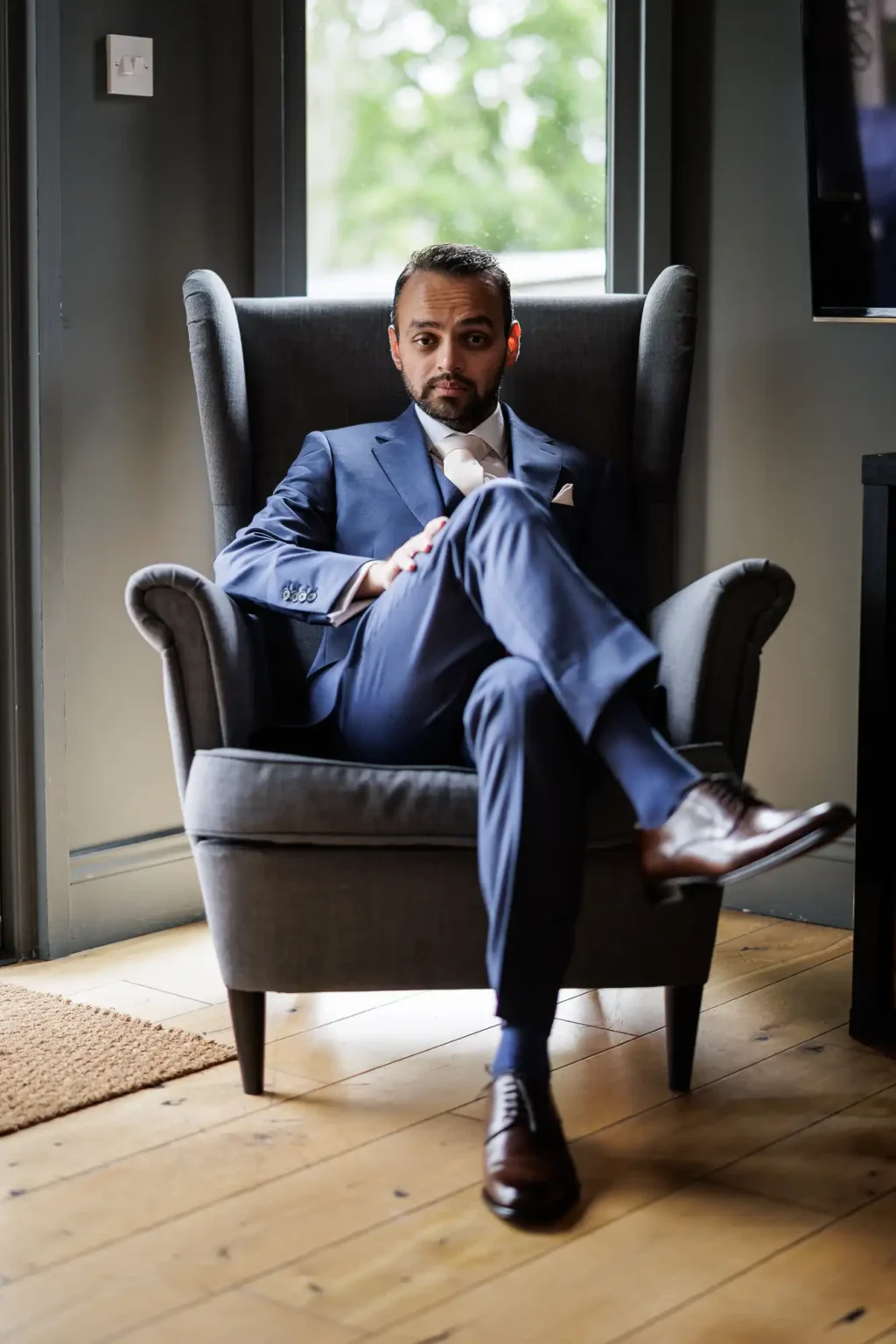 A man in a blue suit sits with legs crossed in a gray armchair, indoors by a window with greenery visible outside.
