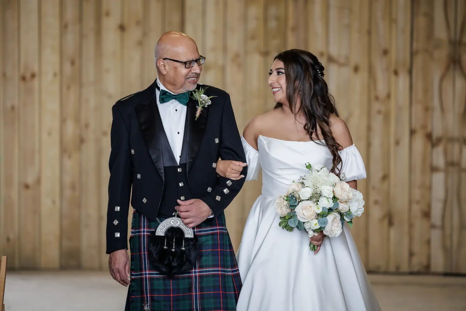 A man in a kilt and tuxedo jacket walks arm-in-arm with a woman in a white wedding dress holding a bouquet, both smiling at each other, in front of a wooden background.