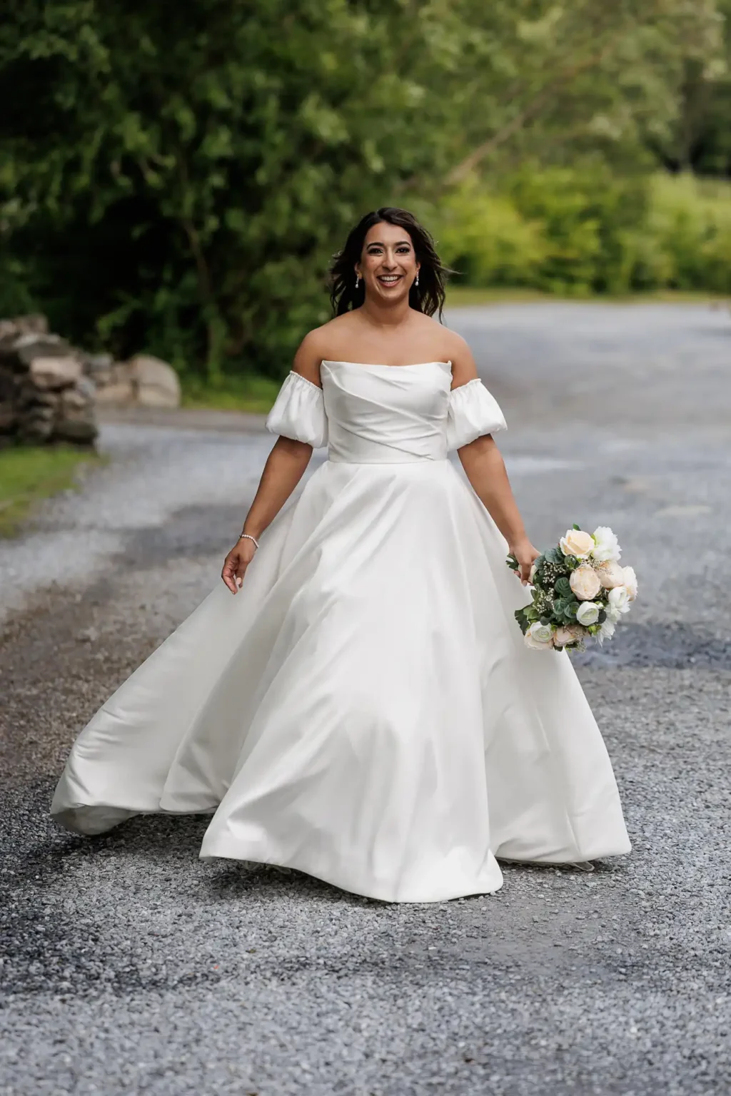 A woman in an off-shoulder white wedding dress holds a bouquet of flowers and smiles while walking on a gravel path outdoors.