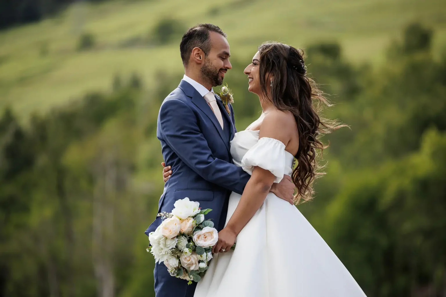A bride and groom stand outdoors, smiling at each other. The bride holds a bouquet of white flowers, and both are dressed in formal wedding attire with a green landscape behind them.