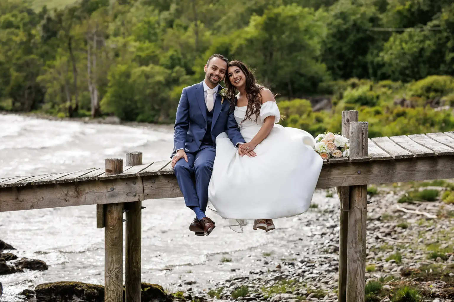 A couple in wedding attire sits together on a wooden dock by a river, surrounded by greenery, with a bouquet of flowers placed beside them.