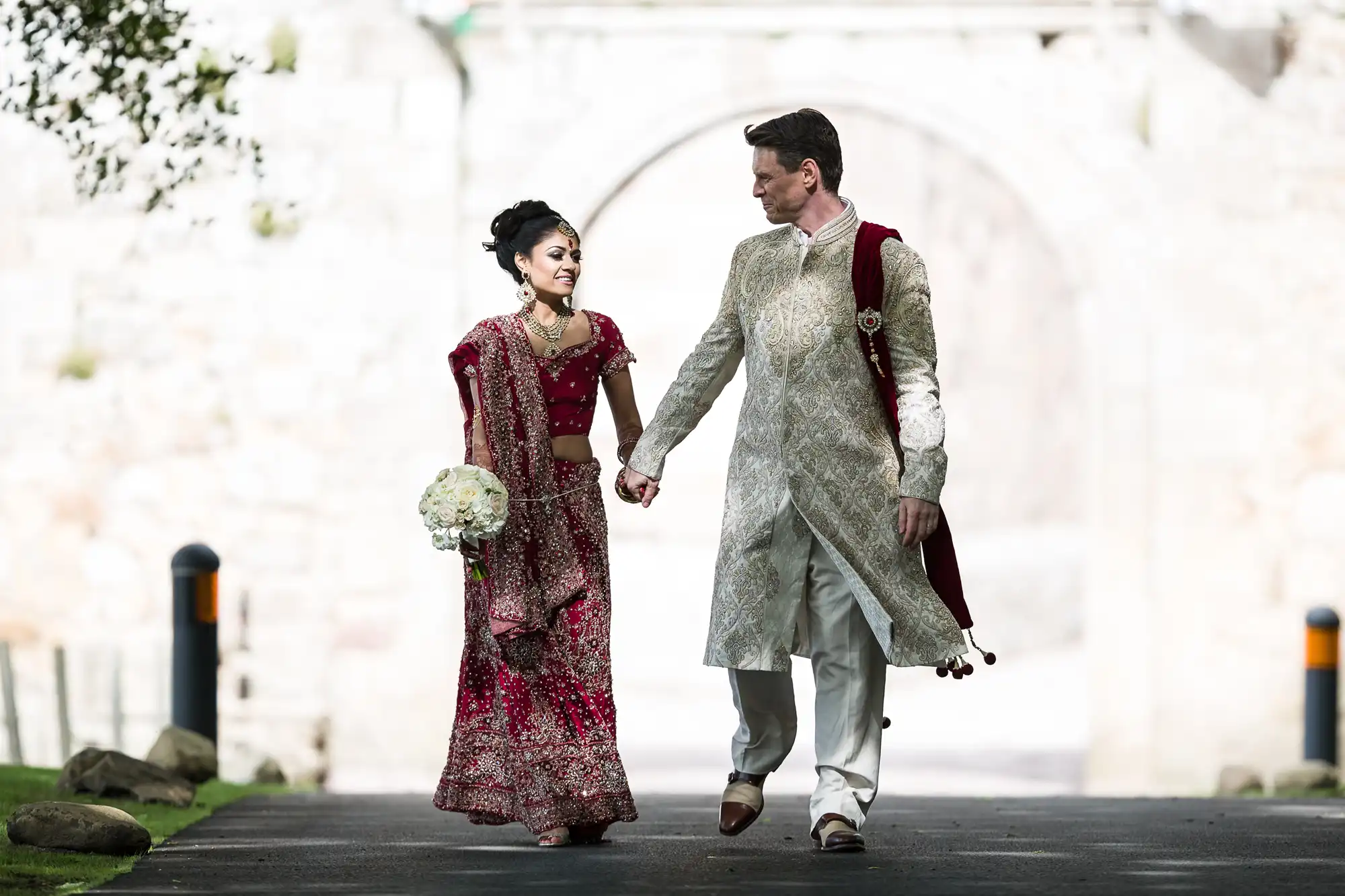 A bride and groom hold hands and walk outdoors. The bride wears a red and gold sari and holds a bouquet. The groom wears a light-colored sherwani. Both appear to be smiling.