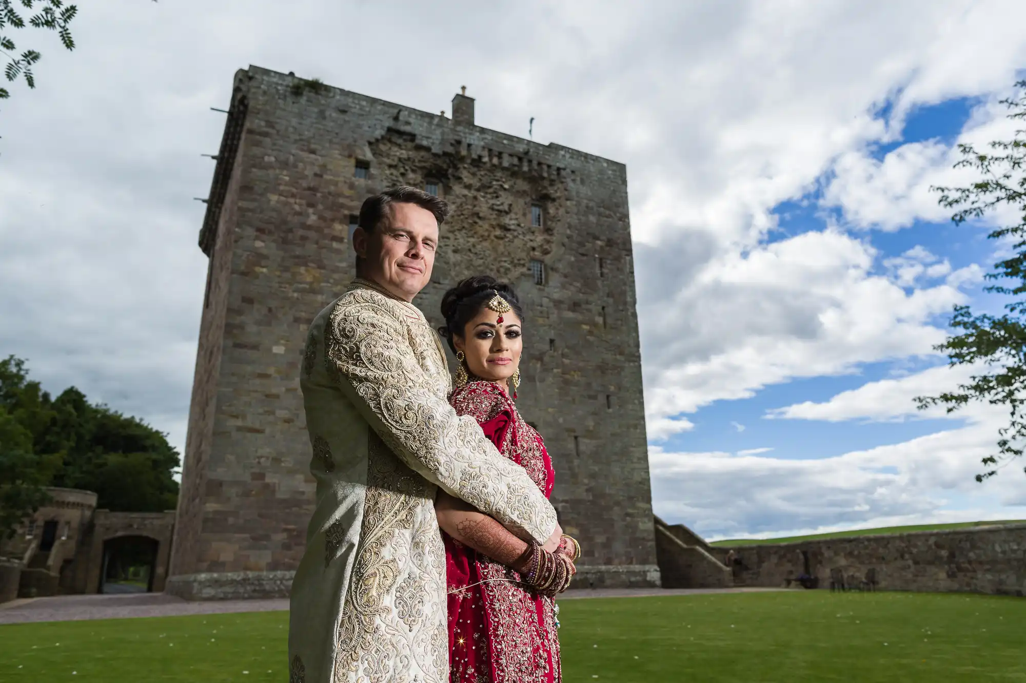 A couple, dressed in traditional attire, stand arm in arm with a historic castle building and a clear sky in the background.
