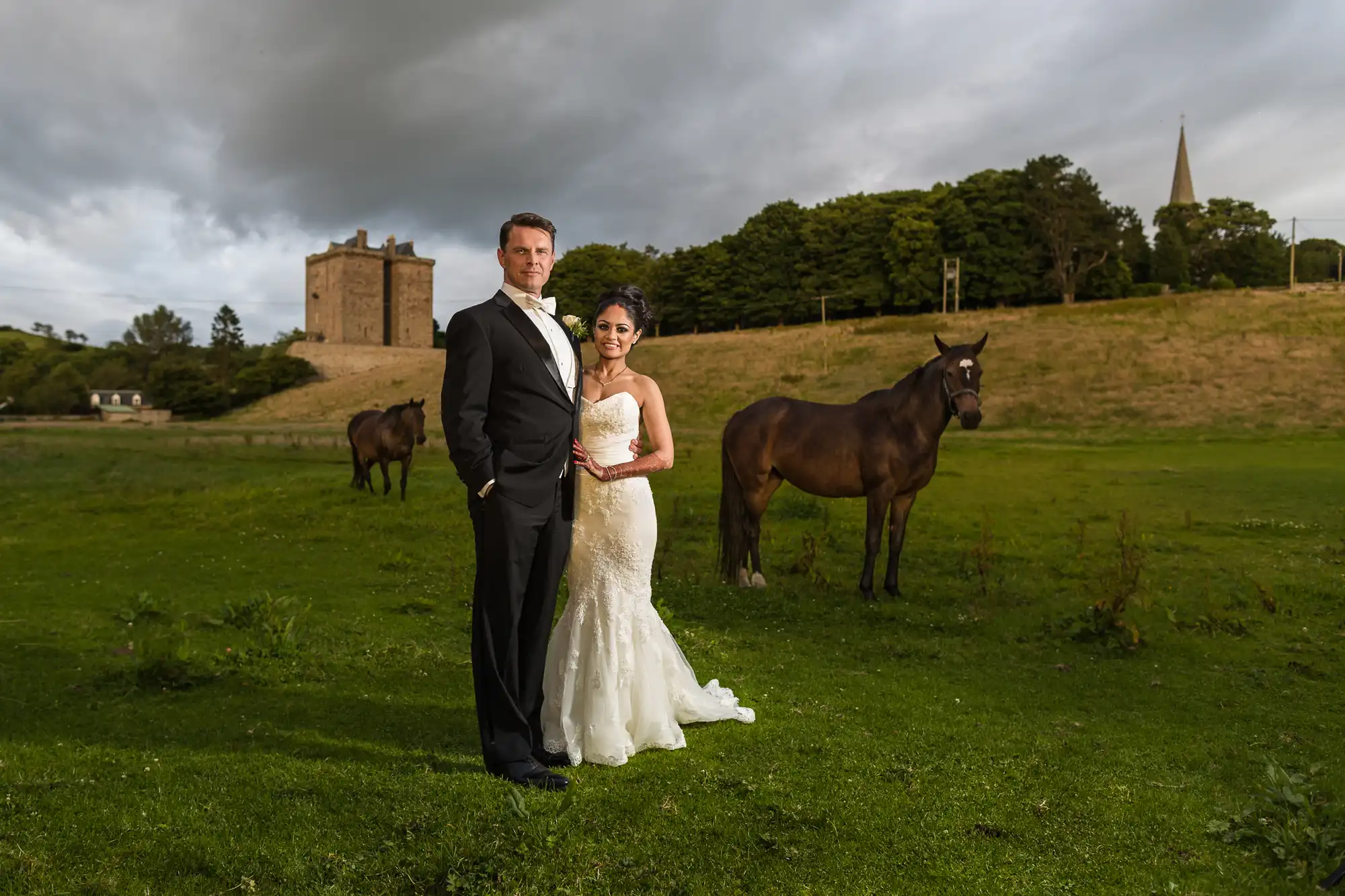 A couple in formal attire stands together on a grassy field with horses nearby and a castle in the background under a cloudy sky.