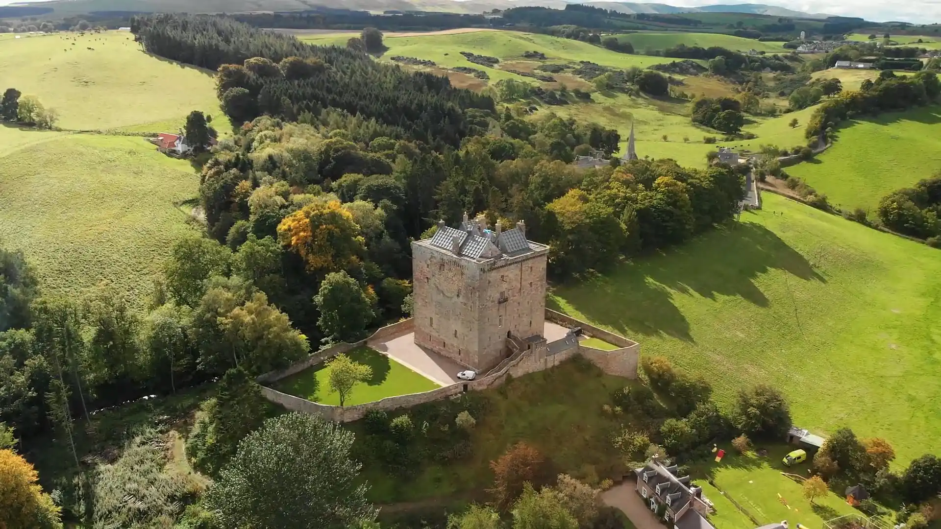 Aerial view of a stone castle surrounded by a wall, set amid green fields and trees in a rural landscape. Borthwick Castle wedding videographer filming Chris and Lee's same-sex marriage celebration - drone shot