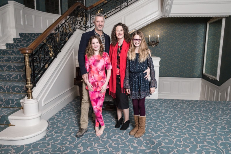 A family of five, including two young girls, posing together on an ornate staircase in a grand room with vintage decor.