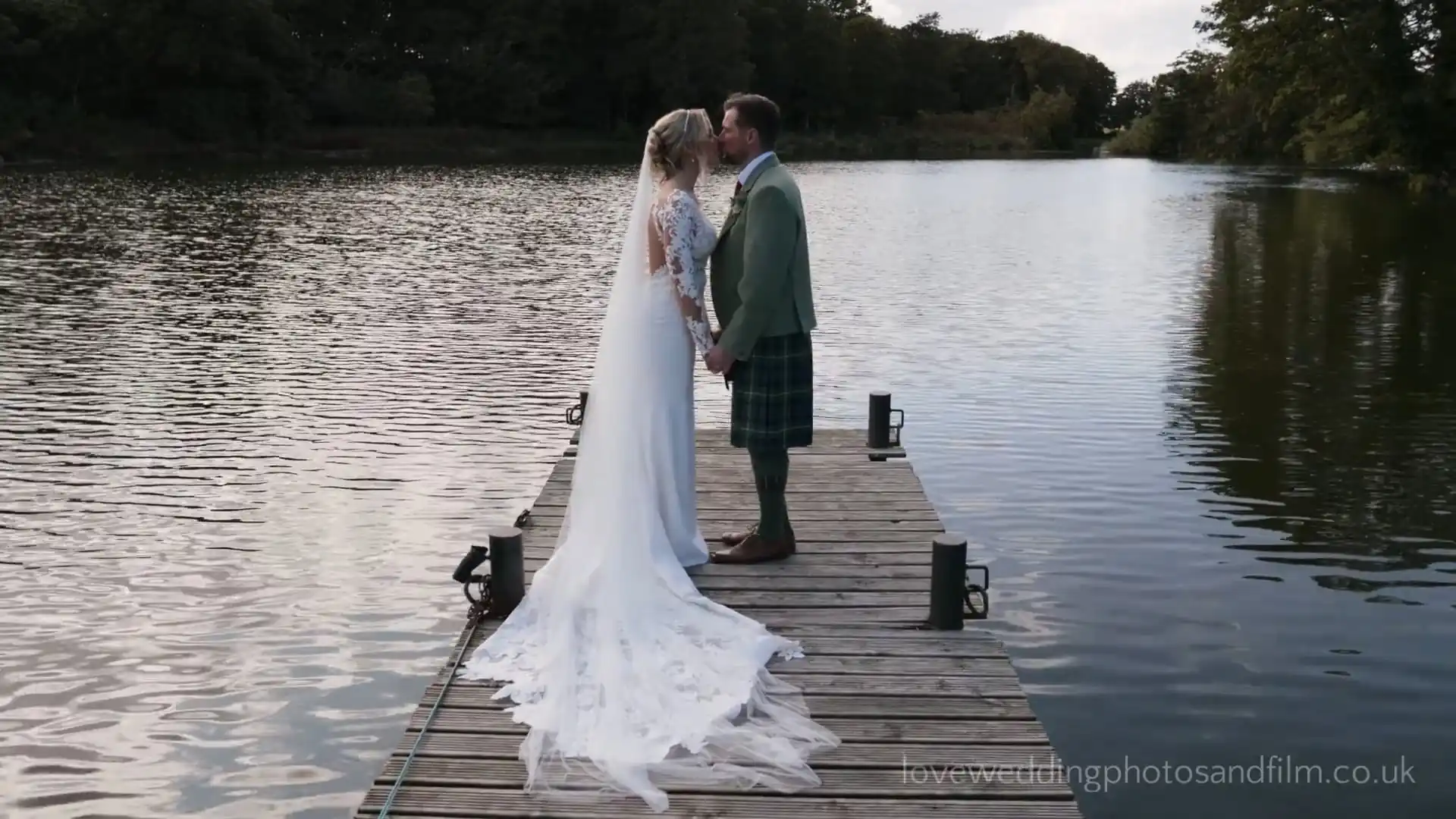 A bride and groom stand on a wooden dock by a lake, holding hands and kissing, surrounded by trees.