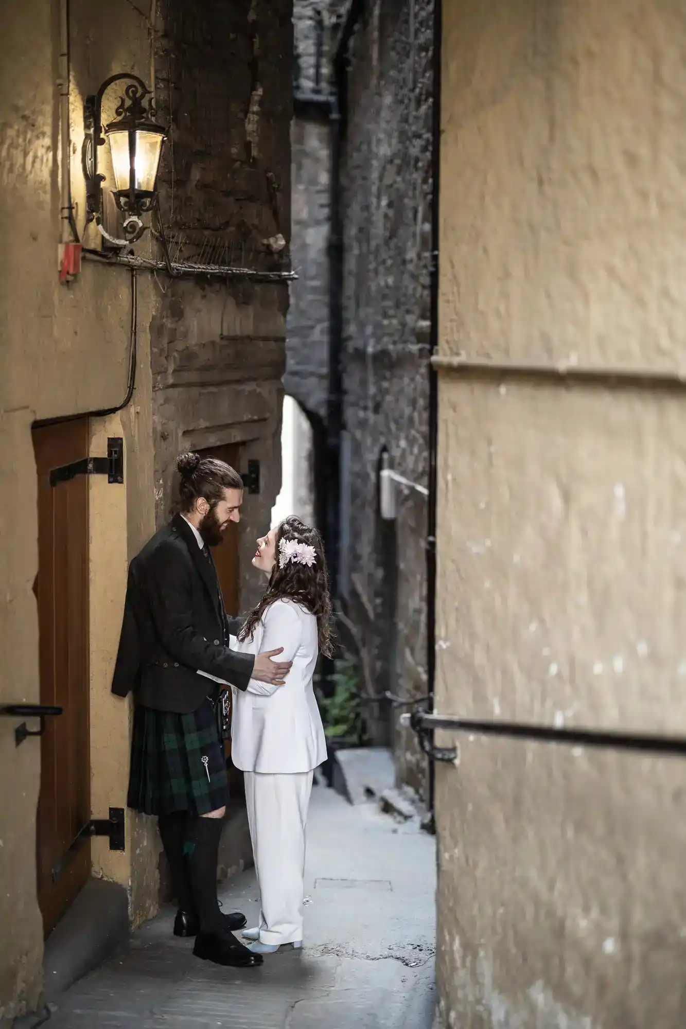 A couple stands facing each other in a narrow alleyway; the man wears a kilt and jacket, and the woman wears a white suit with a floral headpiece.