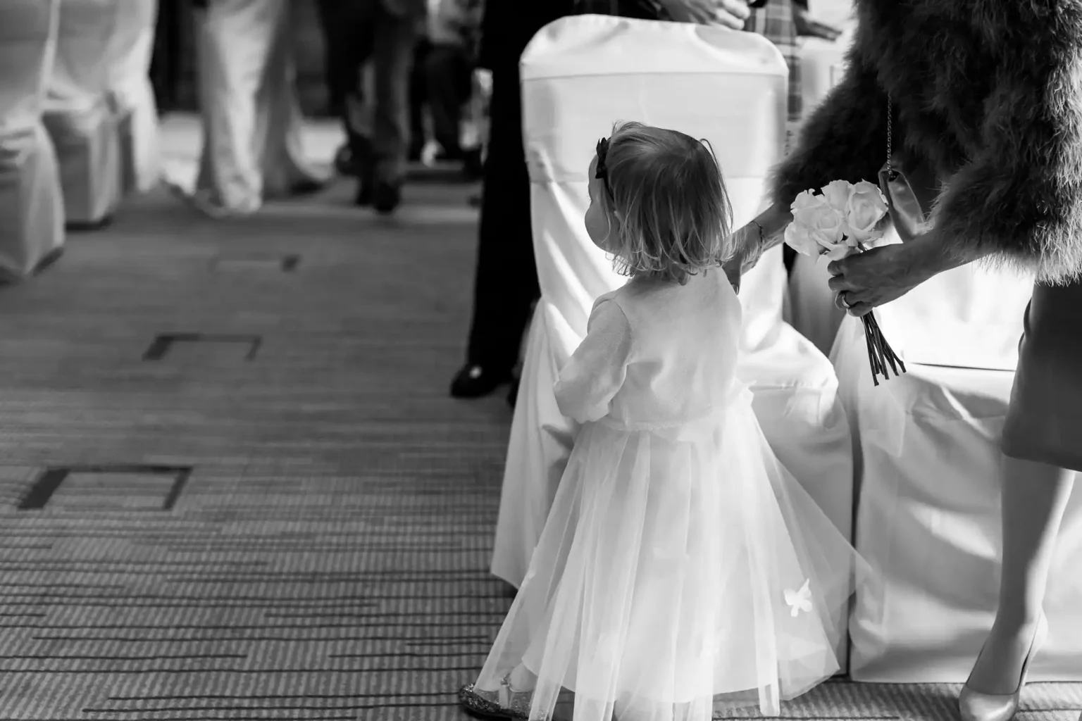 A child in a light dress is standing next to an aisle decorated with white chairs, while an adult in a fur coat reaches over her holding a bouquet of white flowers.