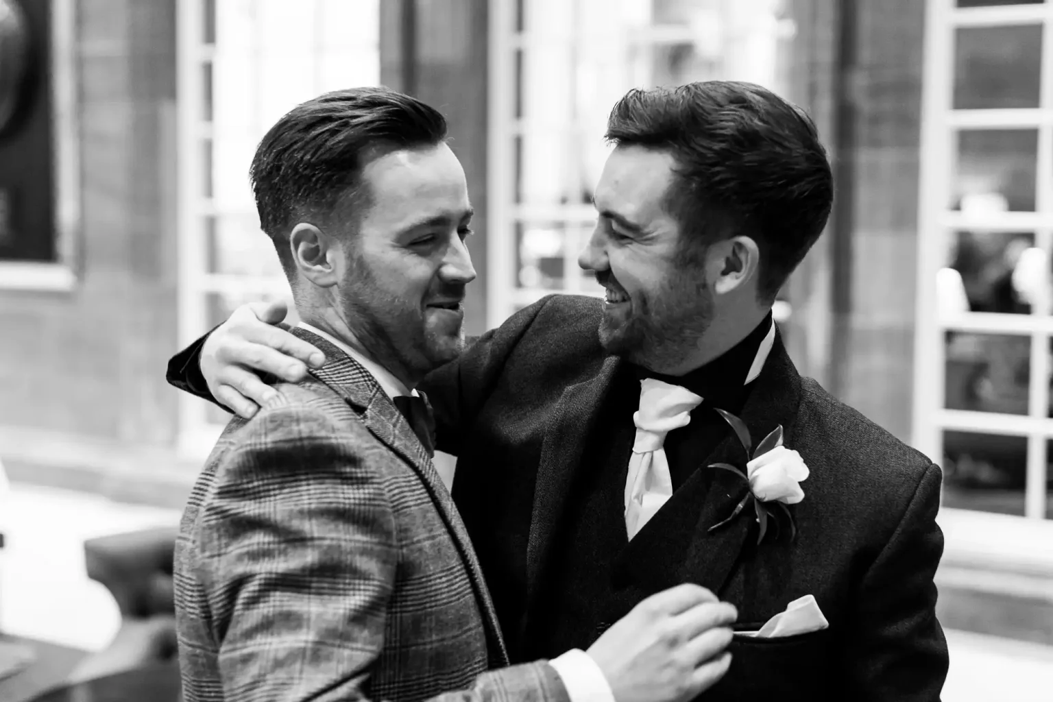 Two men wearing suits, one in a checkered pattern and the other in a solid color, share a moment of laughter and camaraderie in an indoor setting.