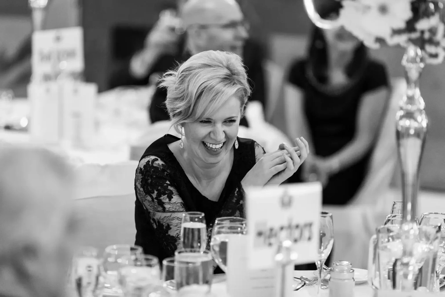 A woman is seated at a decorated table, laughing with others in a formal setting. The table is adorned with glasses and name cards.