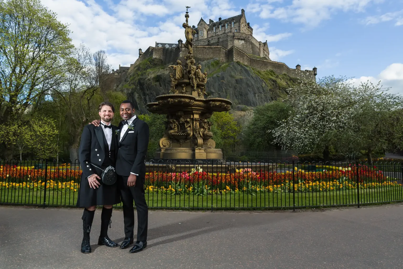 Two grooms, one in a kilt and the other in a suit, stand arm-in-arm in front of a fountain with Edinburgh Castle in the background. Vibrant flowers and trees surround the scene.