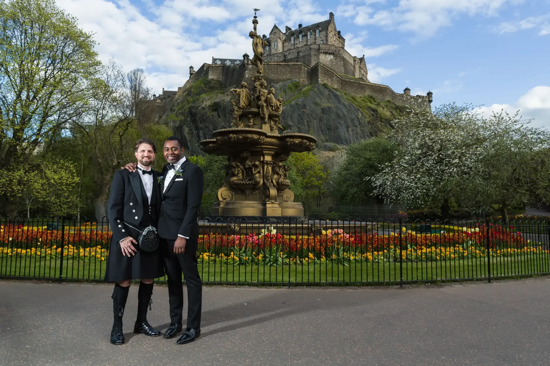 Two grooms, one in a kilt and the other in a suit, stand arm-in-arm in front of a fountain with Edinburgh Castle in the background. Vibrant flowers and trees surround the scene.