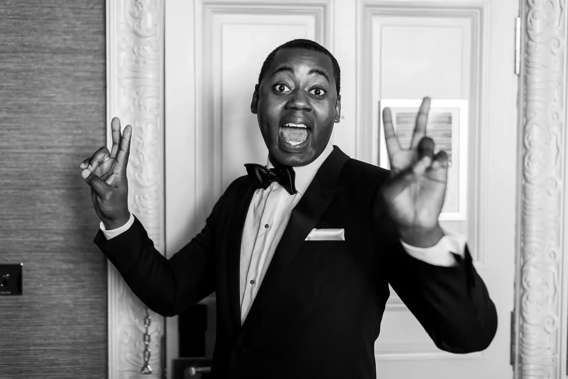 A groom in a tuxedo stands in front of a door, making peace signs with both hands and smiling widely.