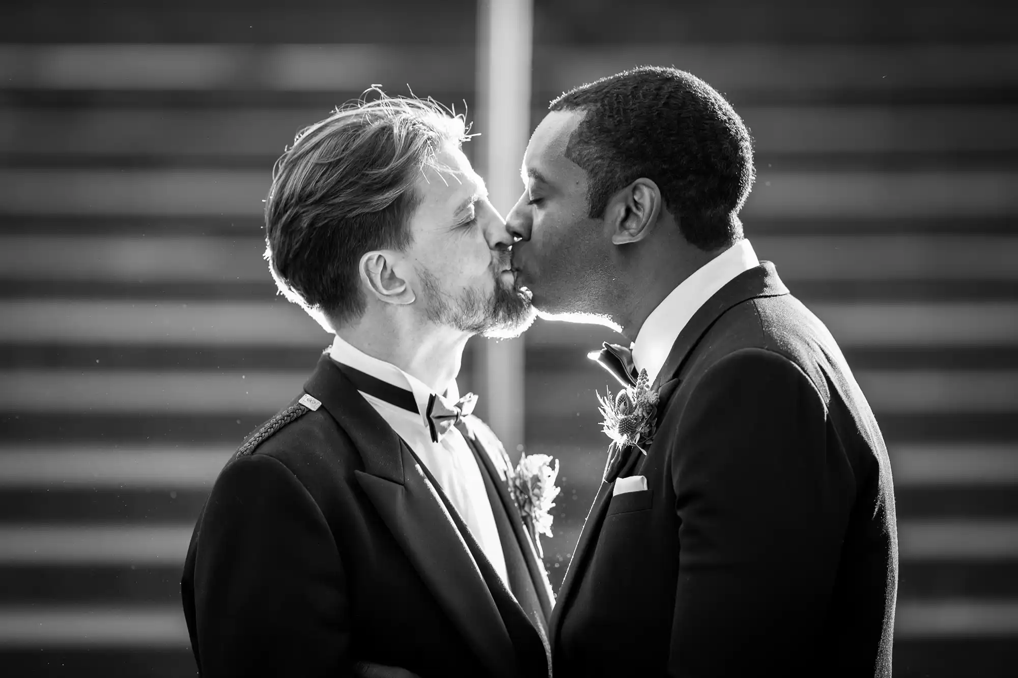 Two men in tuxedos sharing a kiss in front of a staircase. Both are wearing boutonnières.