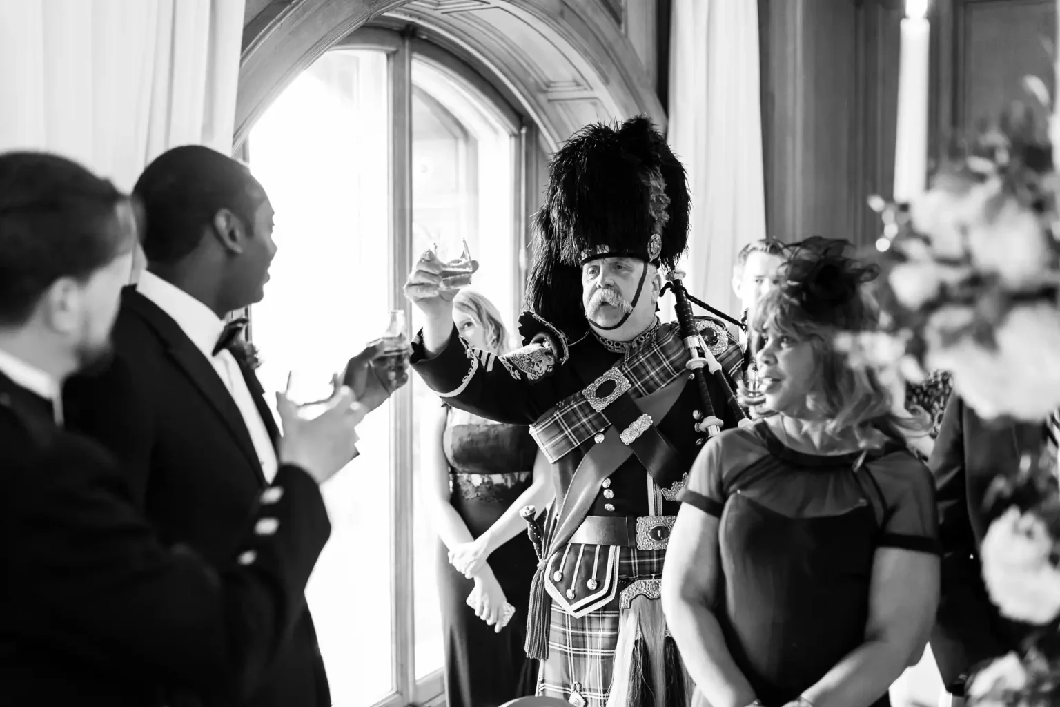 A man in traditional Scottish military attire raises a glass in a toast during a gathering. Several attendees in formal outfits are watching him and also raising their glasses.