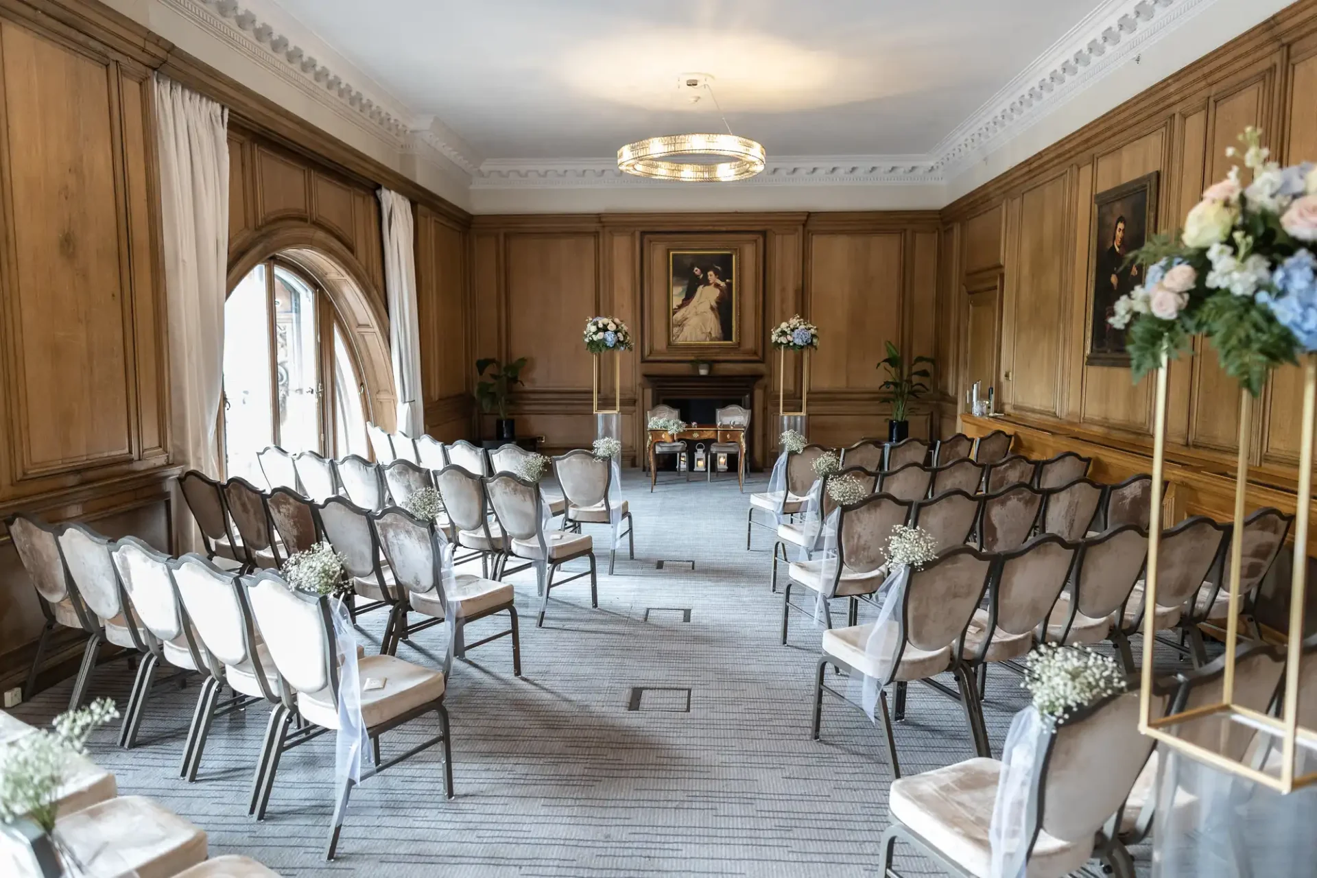 Caledonian Hotel wedding photos: Elegant interior of a room set up for a small event, with rows of chairs facing a central aisle and floral decorations, wooden paneling, and portrait paintings on the walls.