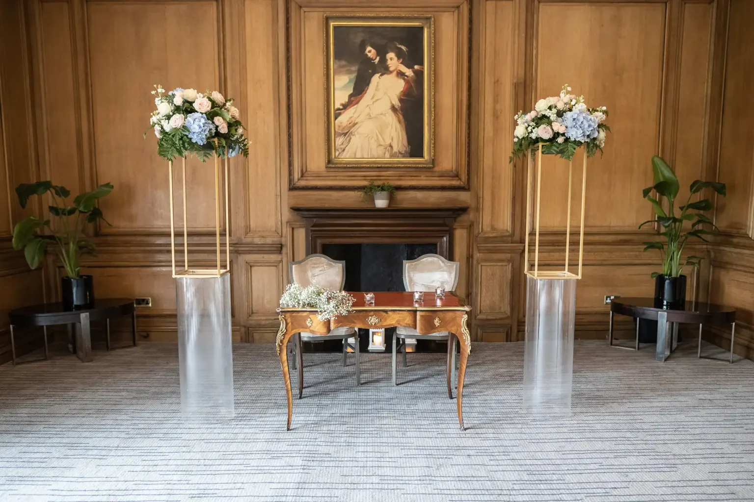 Elegant interior of a room with a vintage painting above a fireplace, flanked by two floral arrangements on tall stands, and a decorative table with two chairs.