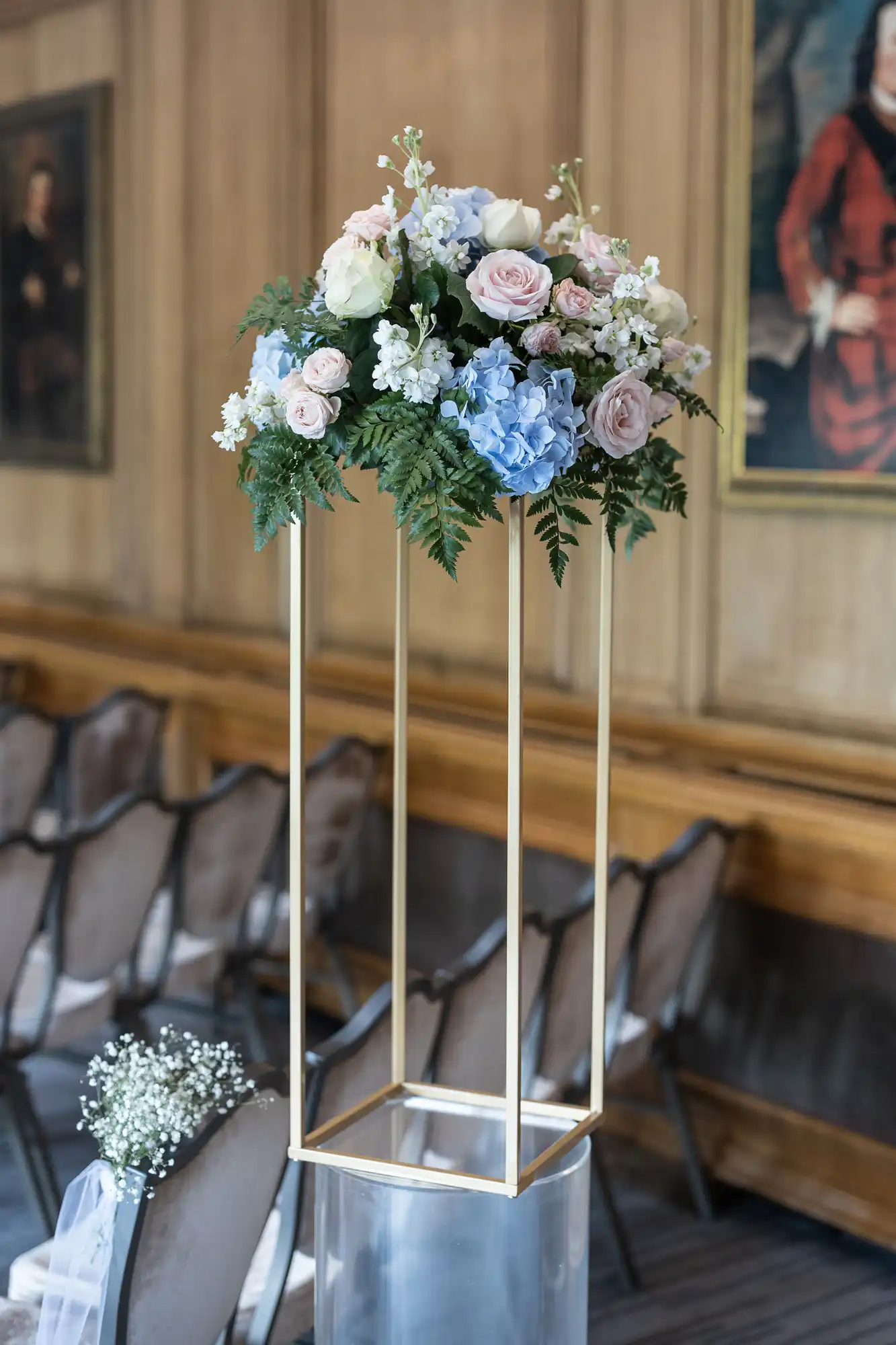 An elegant floral arrangement with pastel roses and hydrangeas atop a tall, gold geometric stand in a vintage room with wooden panels and classic paintings.